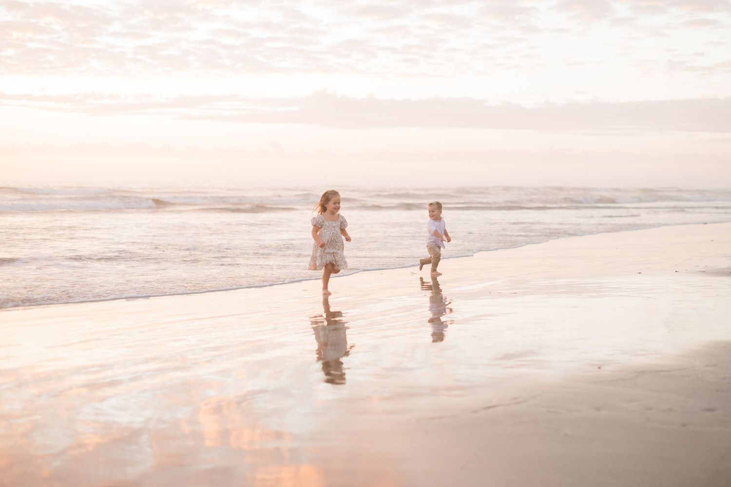 Two children running joyfully along a sandy beach at sunset, waves gently lapping the shore in soft pink light.