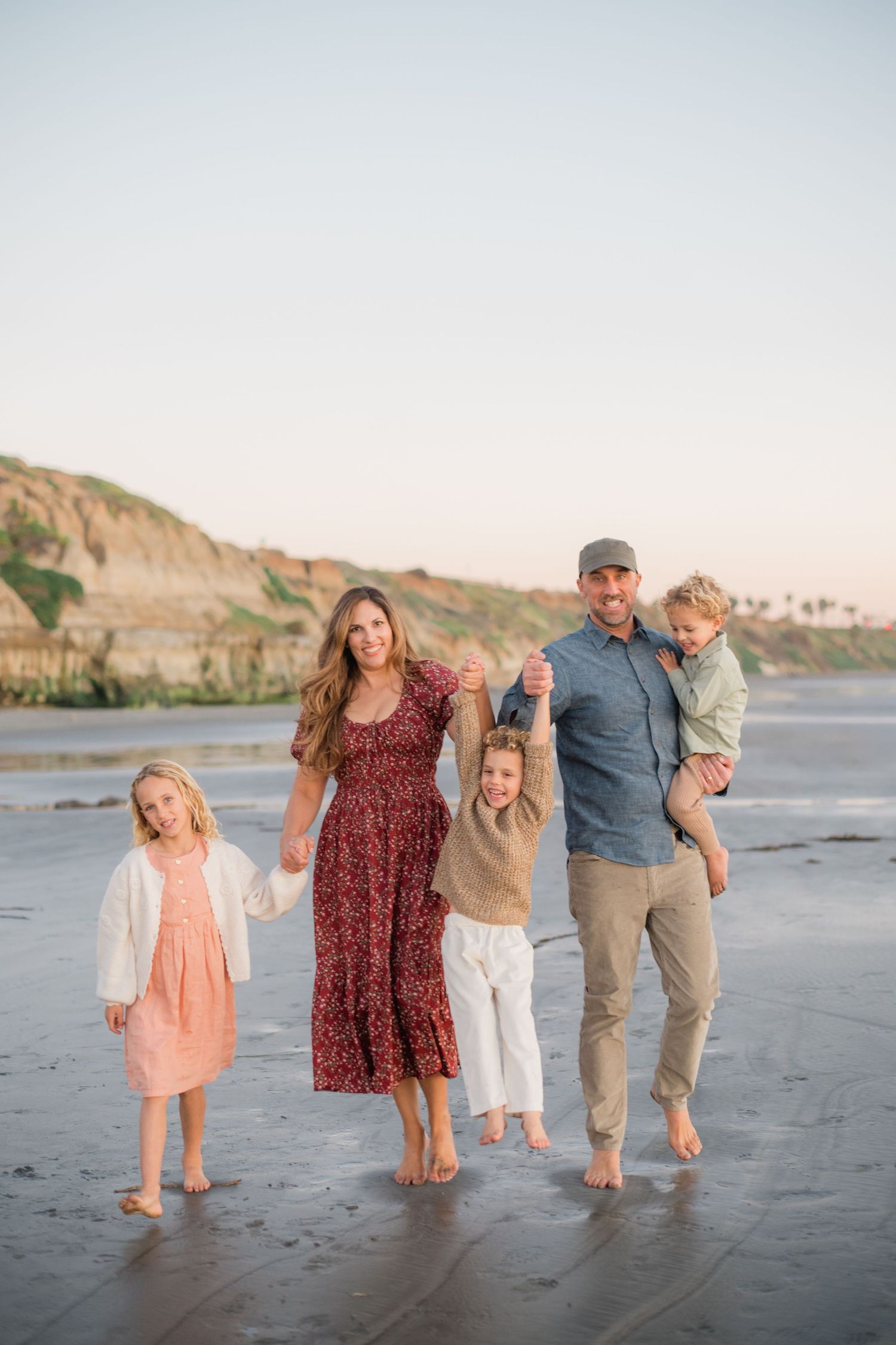 A happy family of five walks barefoot on a beach at sunset with scenic cliffs in the background.