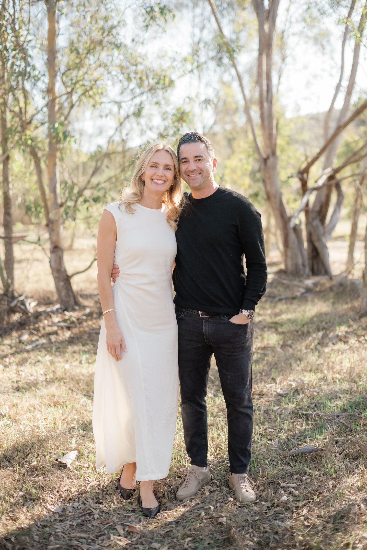 Smiling couple posing outdoors among eucalyptus trees, woman in white dress and man in black sweater.
