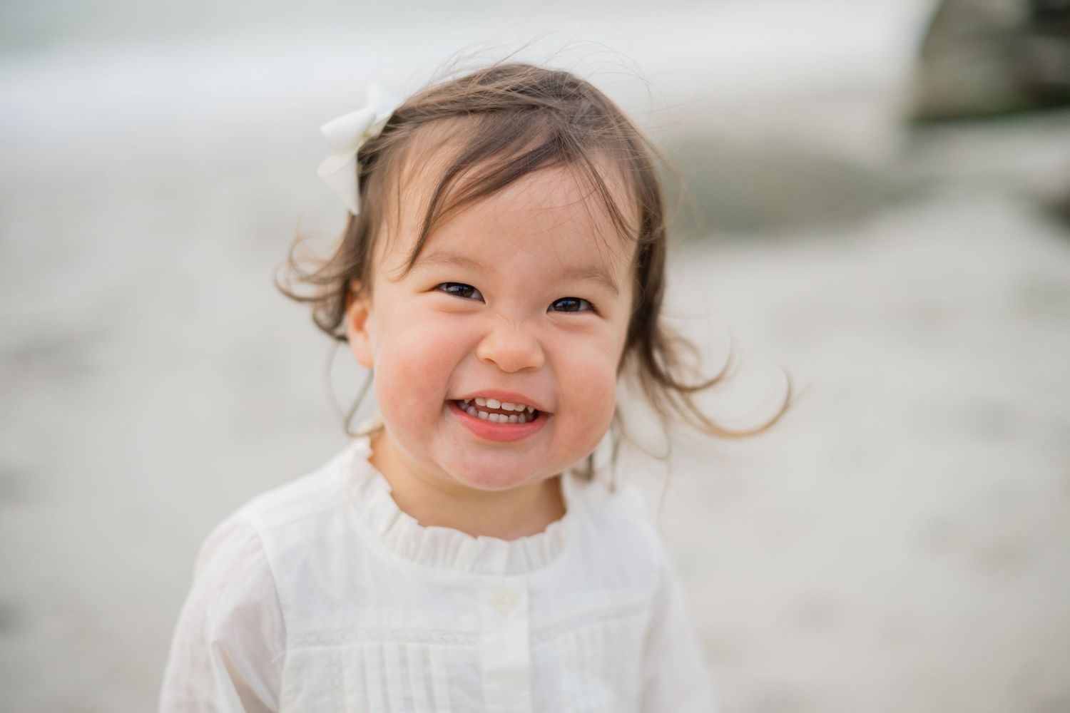 Smiling toddler girl with brown hair wearing a white dress on a beach.