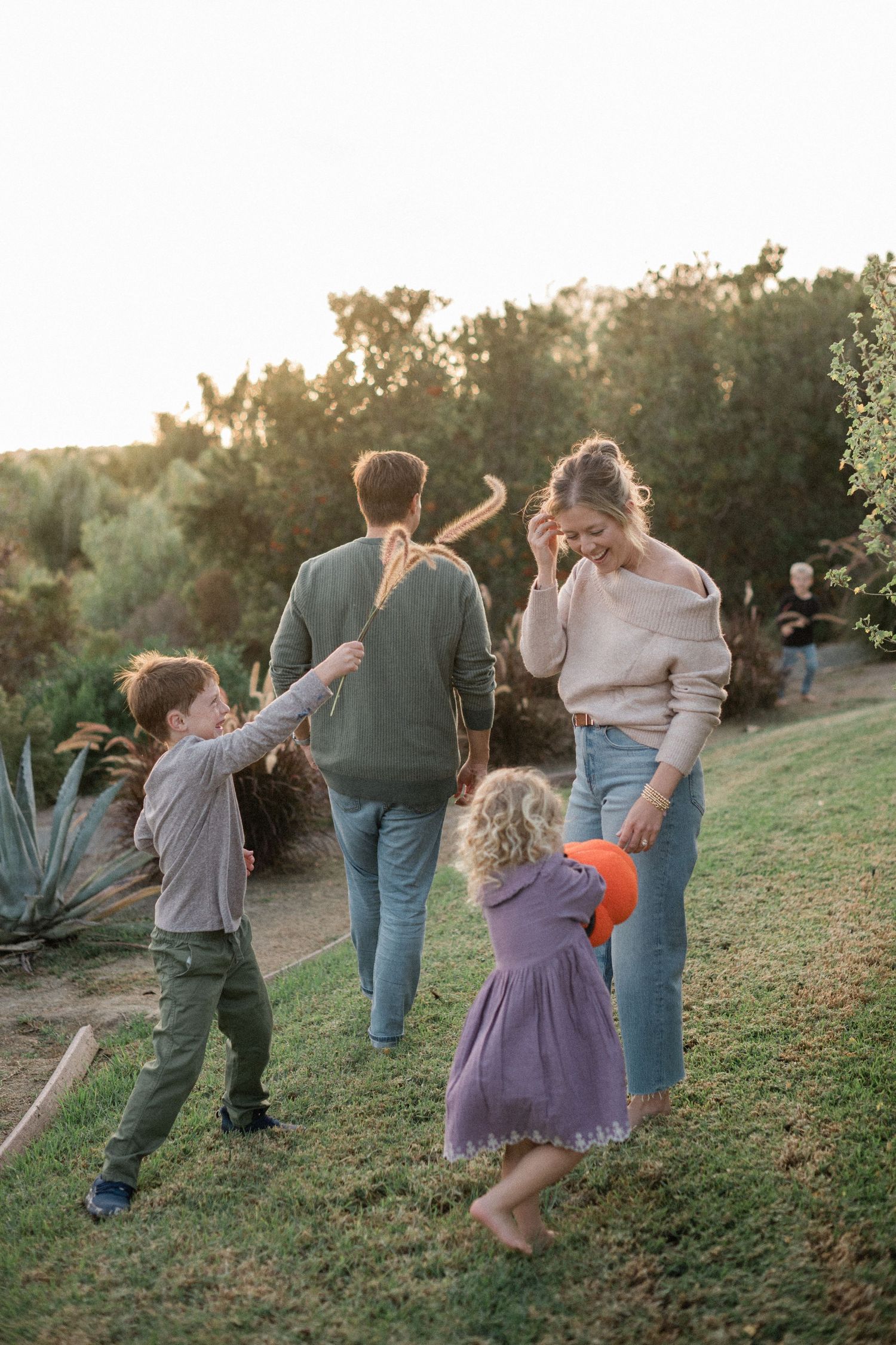 Family of four playing outdoors on green grass at golden hour, with trees and warm sunlight in the background.