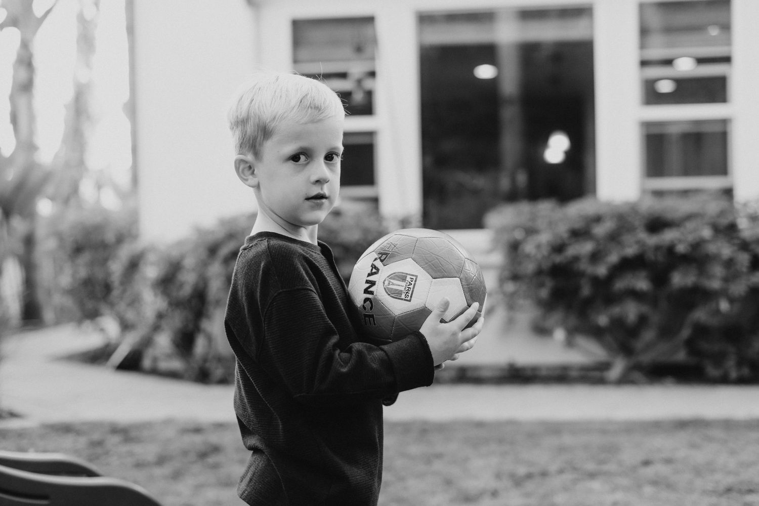 Black and white photo of a young blonde boy holding a soccer ball outdoors near a house.