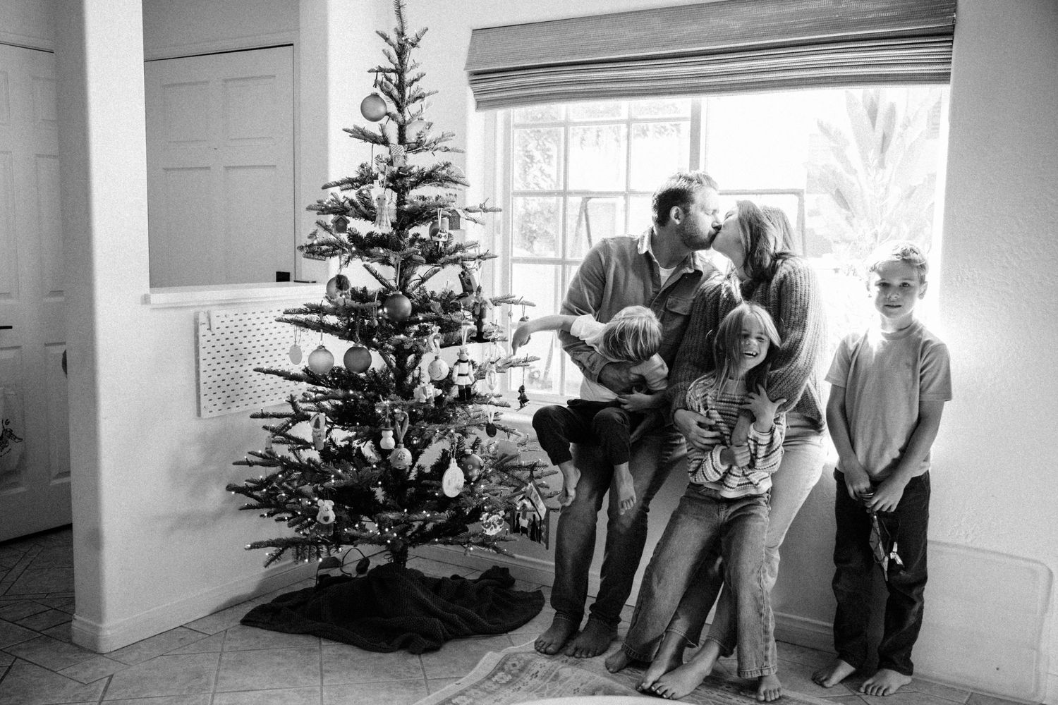 Black and white family Christmas photo with parents kissing beside decorated tree, surrounded by three young children.
