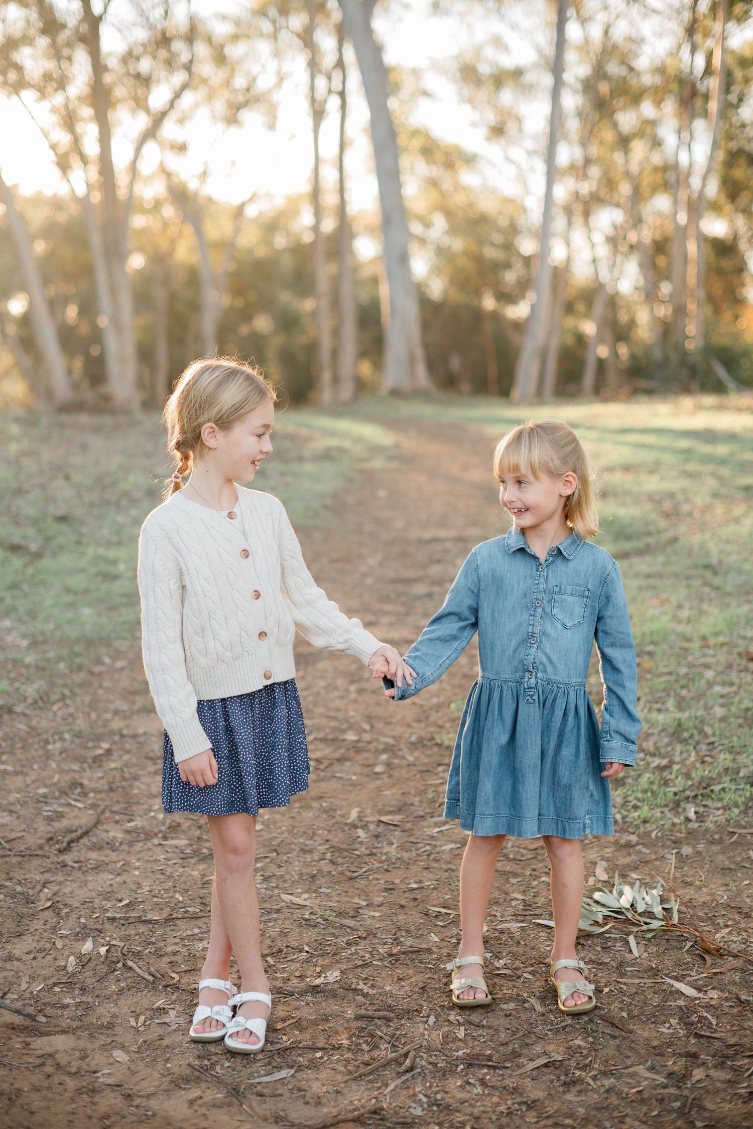 Two young girls in dresses hold hands on a dirt path surrounded by trees in golden hour light.