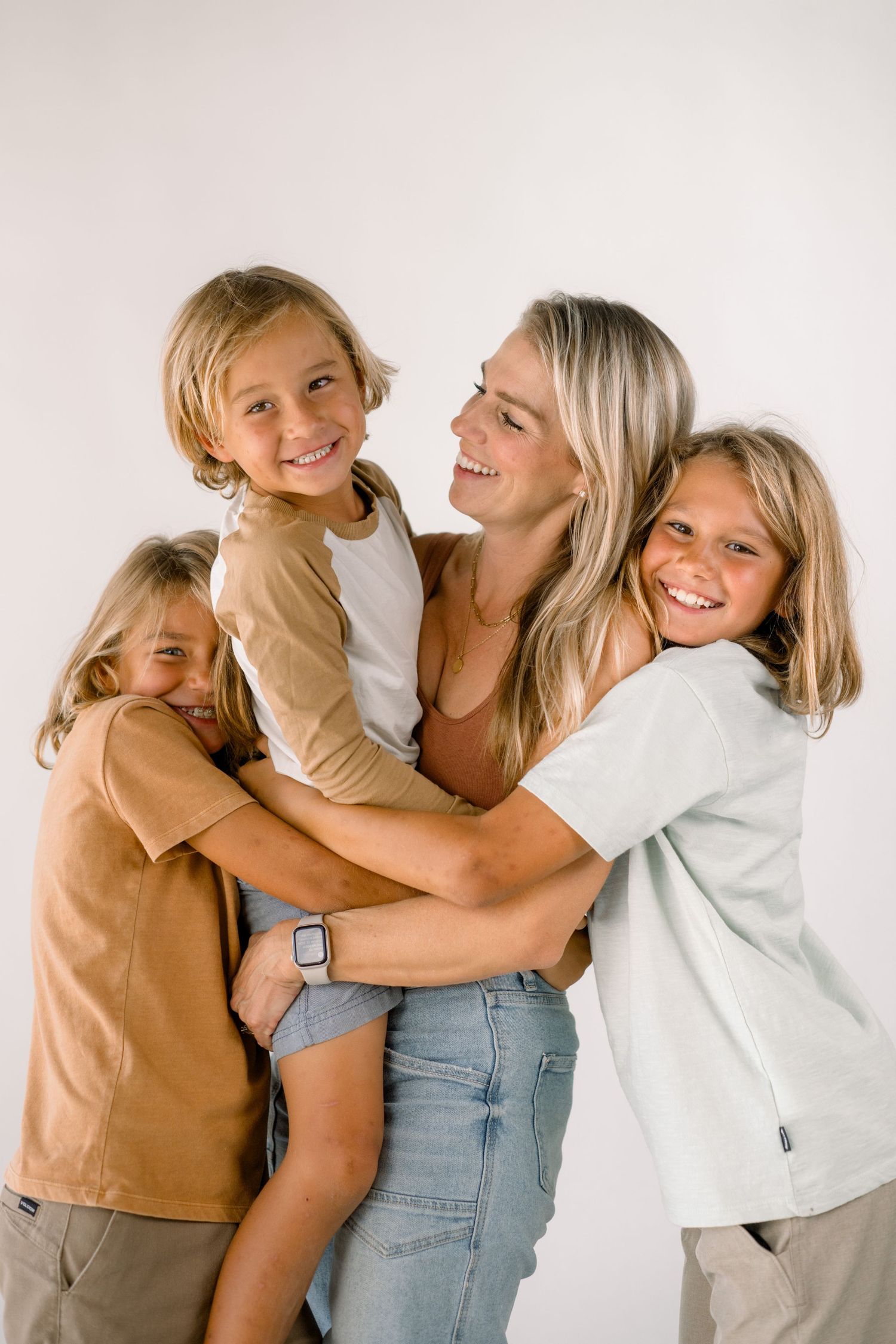 A smiling blonde mother hugs her three happy children in a bright, casual studio family portrait.