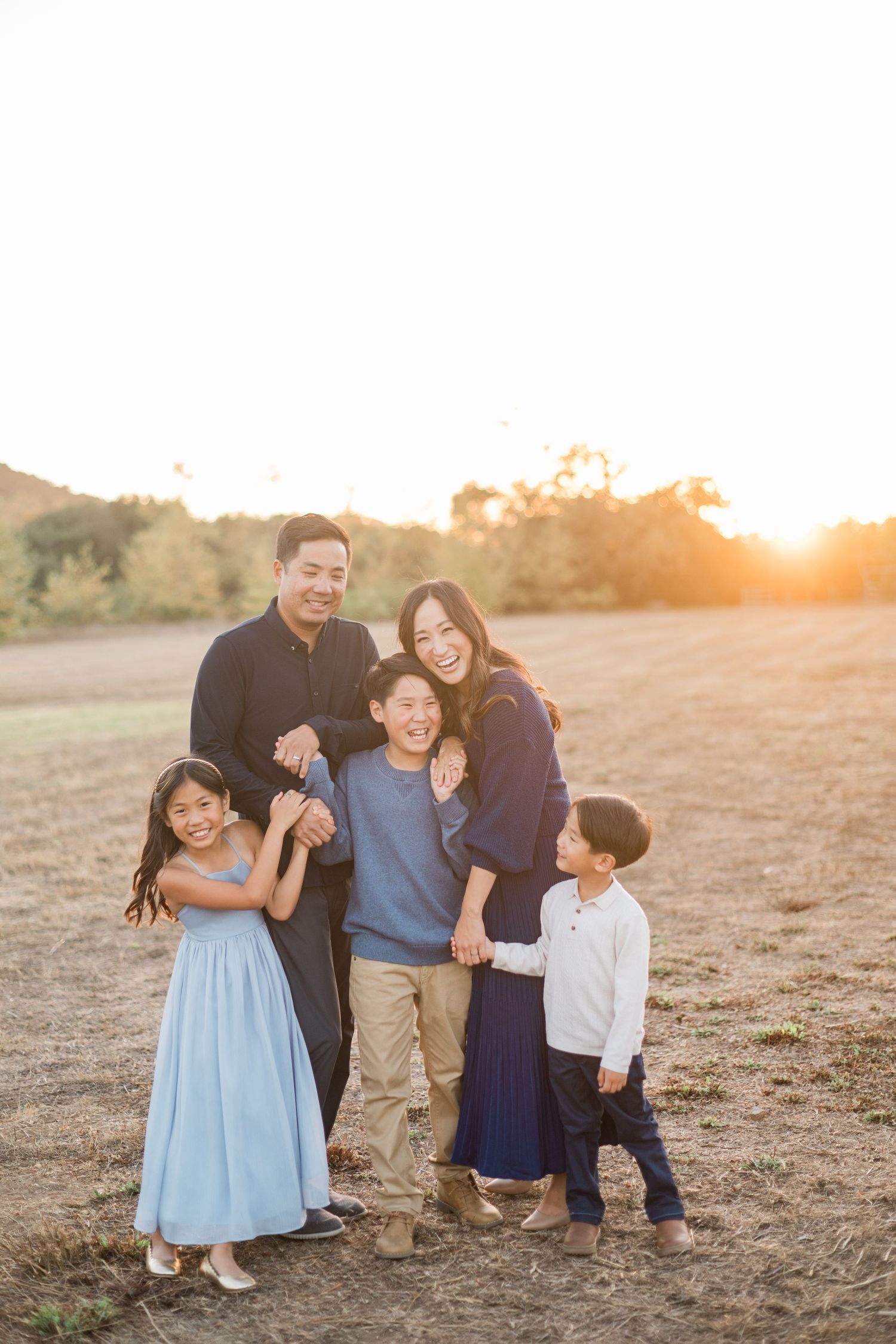 A joyful Asian family of five shares a warm embrace in an open field bathed in beautiful sunset light.