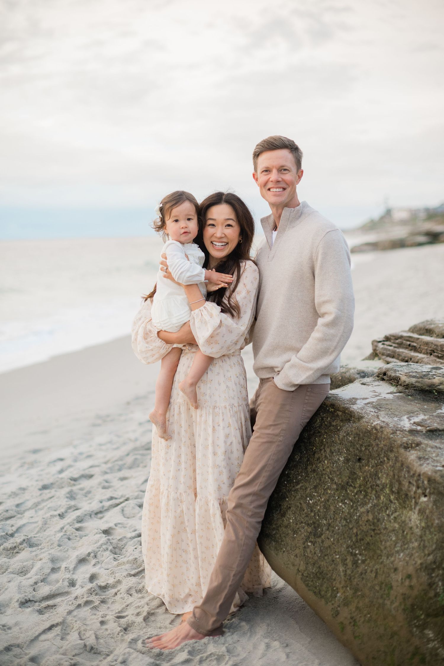 Happy family of three poses on sandy beach, mother holding toddler while father smiles beside her.