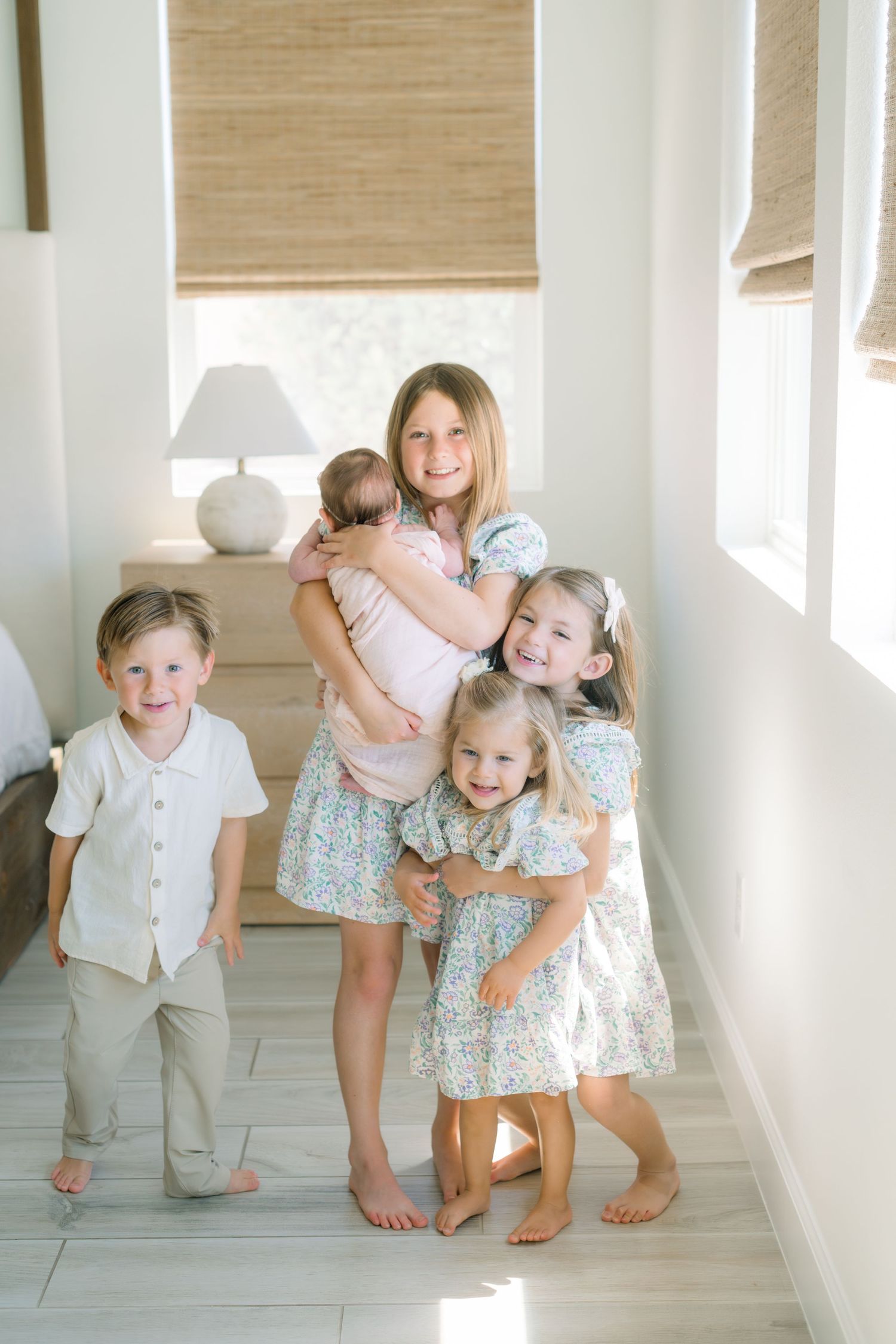 Five young children pose together in a bright, airy room, with the oldest girl hugging a newborn baby.