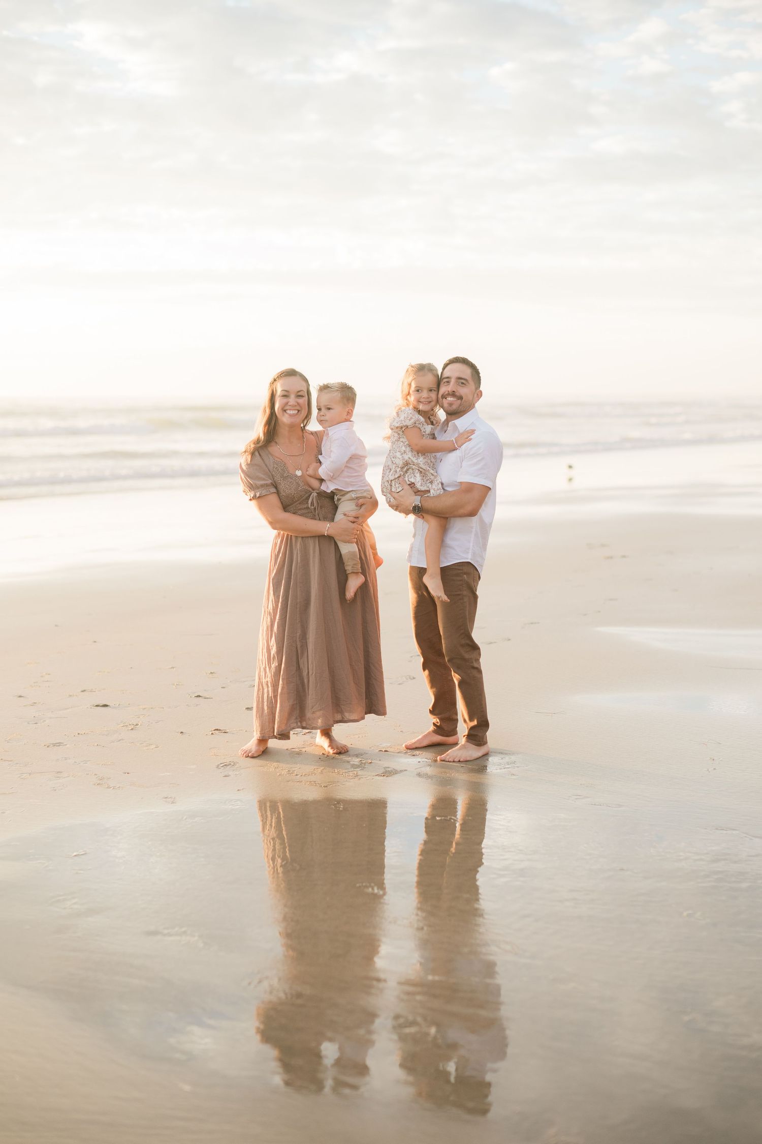 Family of four posing on a sunny beach, parents holding two young children, with ocean waves and reflections in the sand.