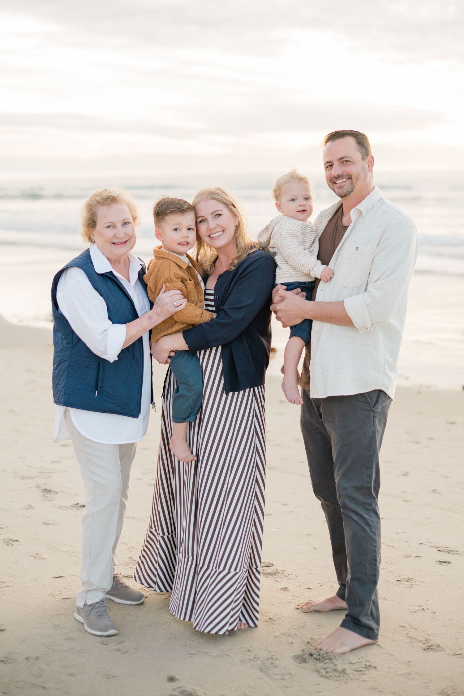 Multi-generational family of five smiling together on a sandy beach at sunset, including grandmother, parents, and two young boys.