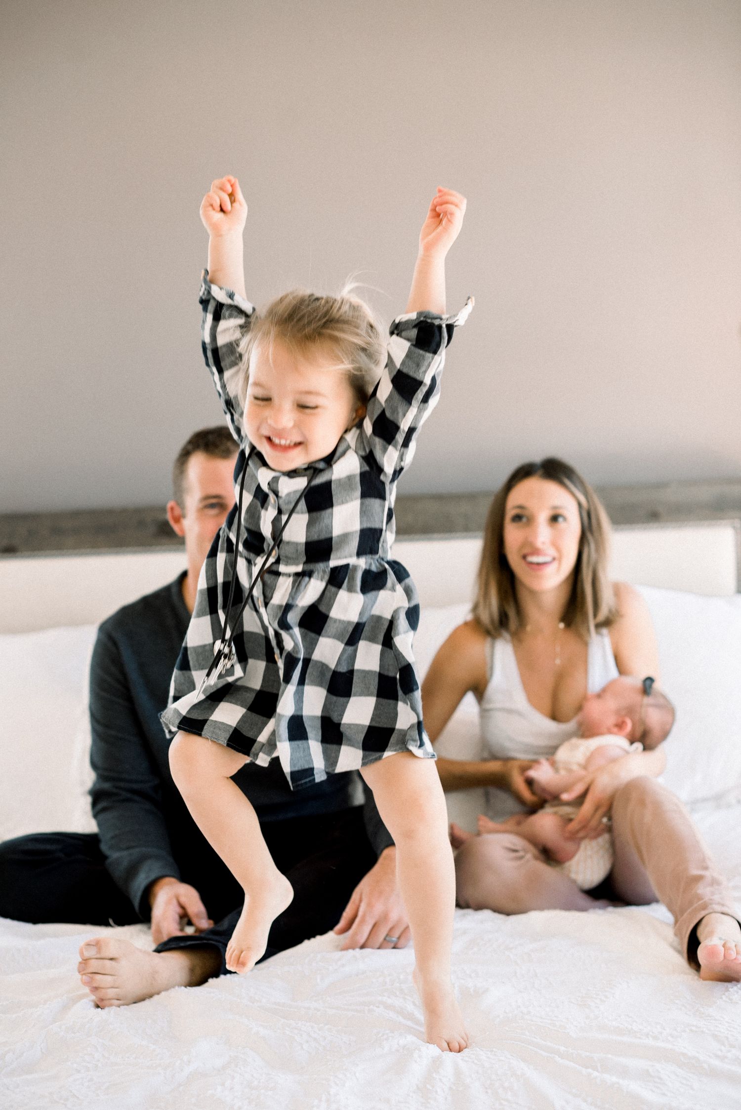A joyful toddler in plaid jumps on a bed while parents sit behind holding a newborn baby.