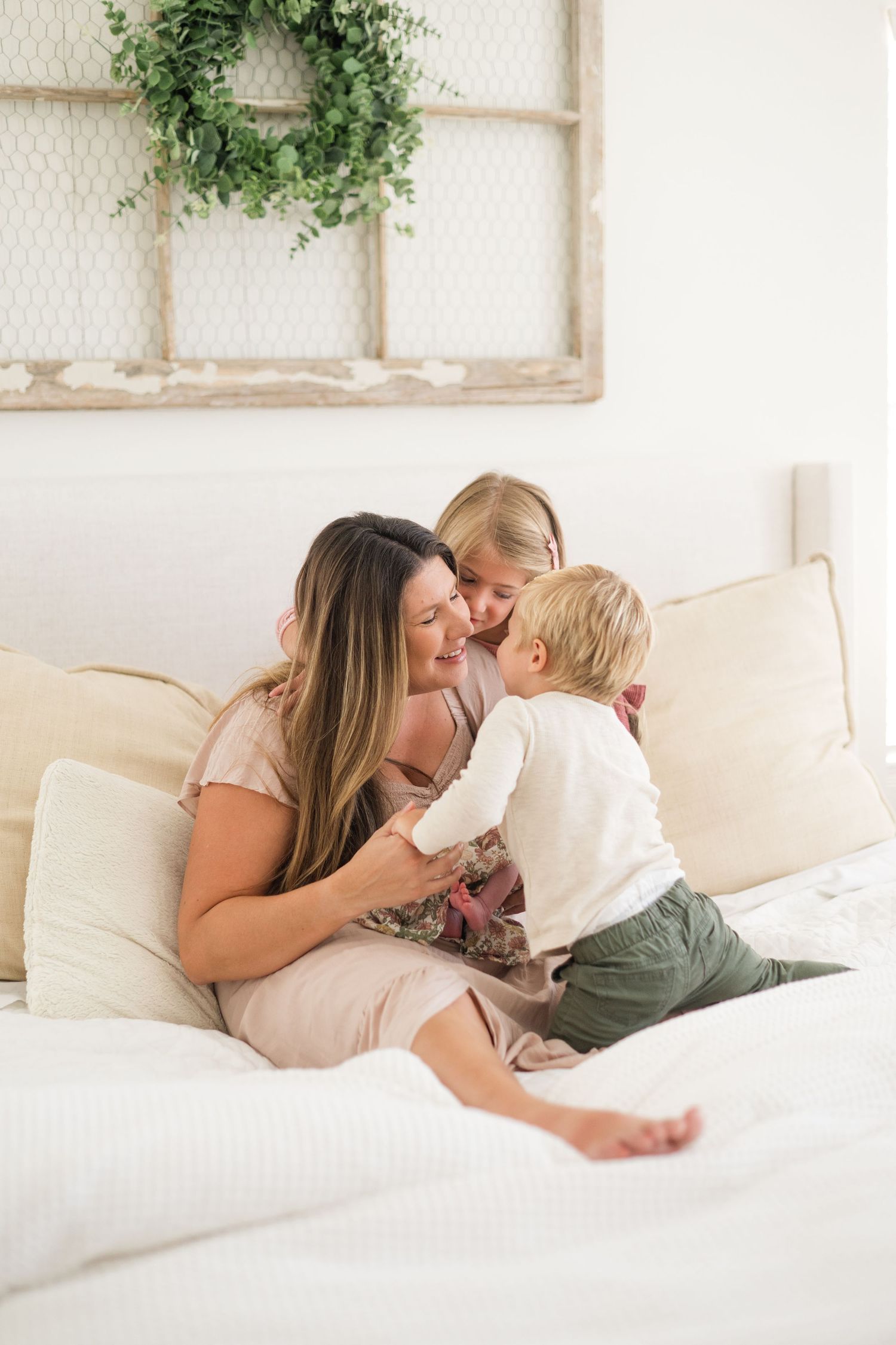 Mother sitting on white bed sharing tender moment with young daughter and toddler son in bright room.
