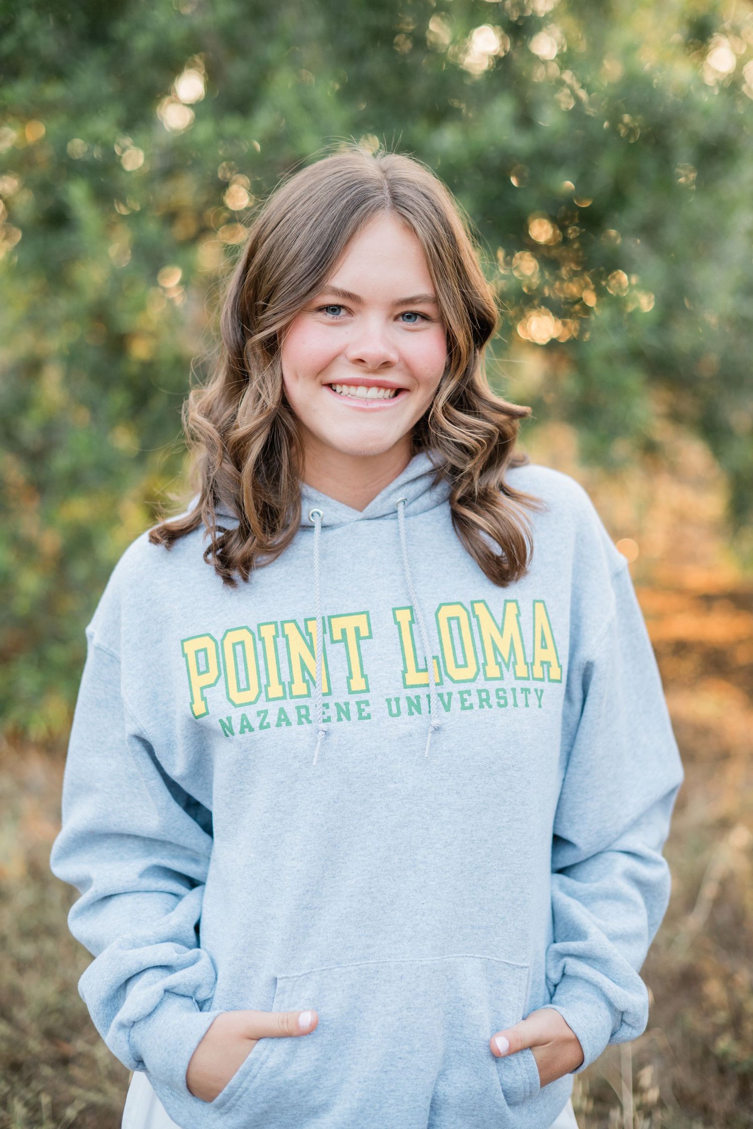 Smiling teen in Point Loma Nazarene University hoodie posing outdoors among green trees.