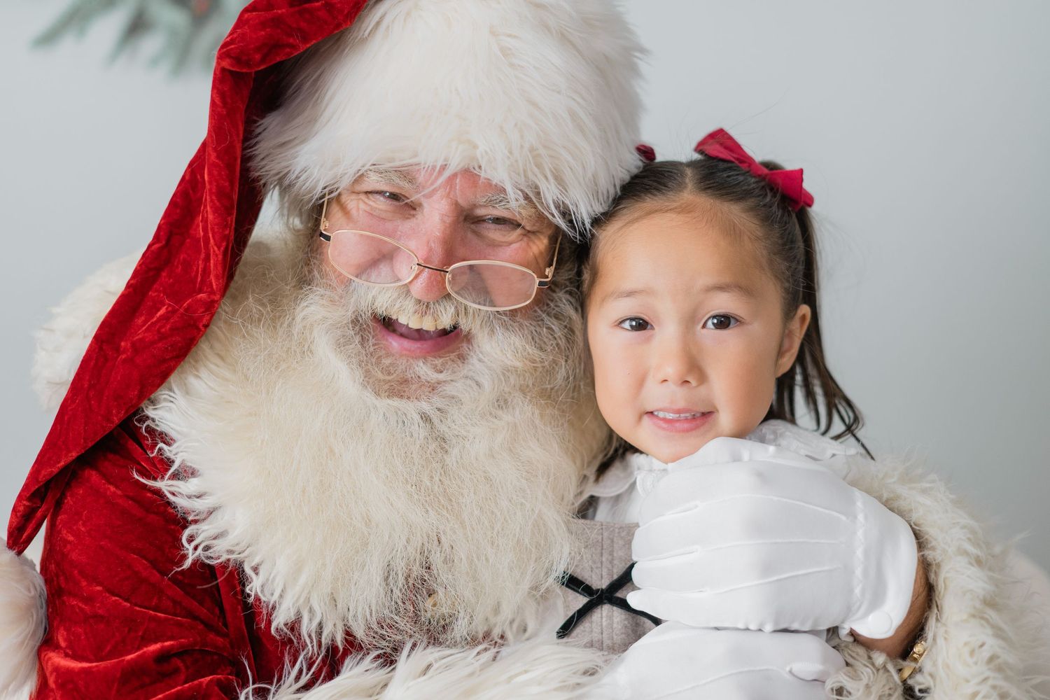 A cheerful Santa Claus poses with a smiling young girl in a white dress with red bow during a Christmas photo session.