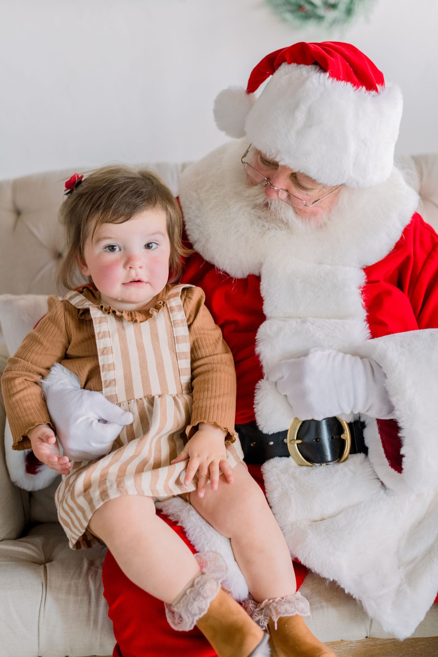Toddler girl in striped dress sitting on Santa Claus lap during a Christmas portrait session.
