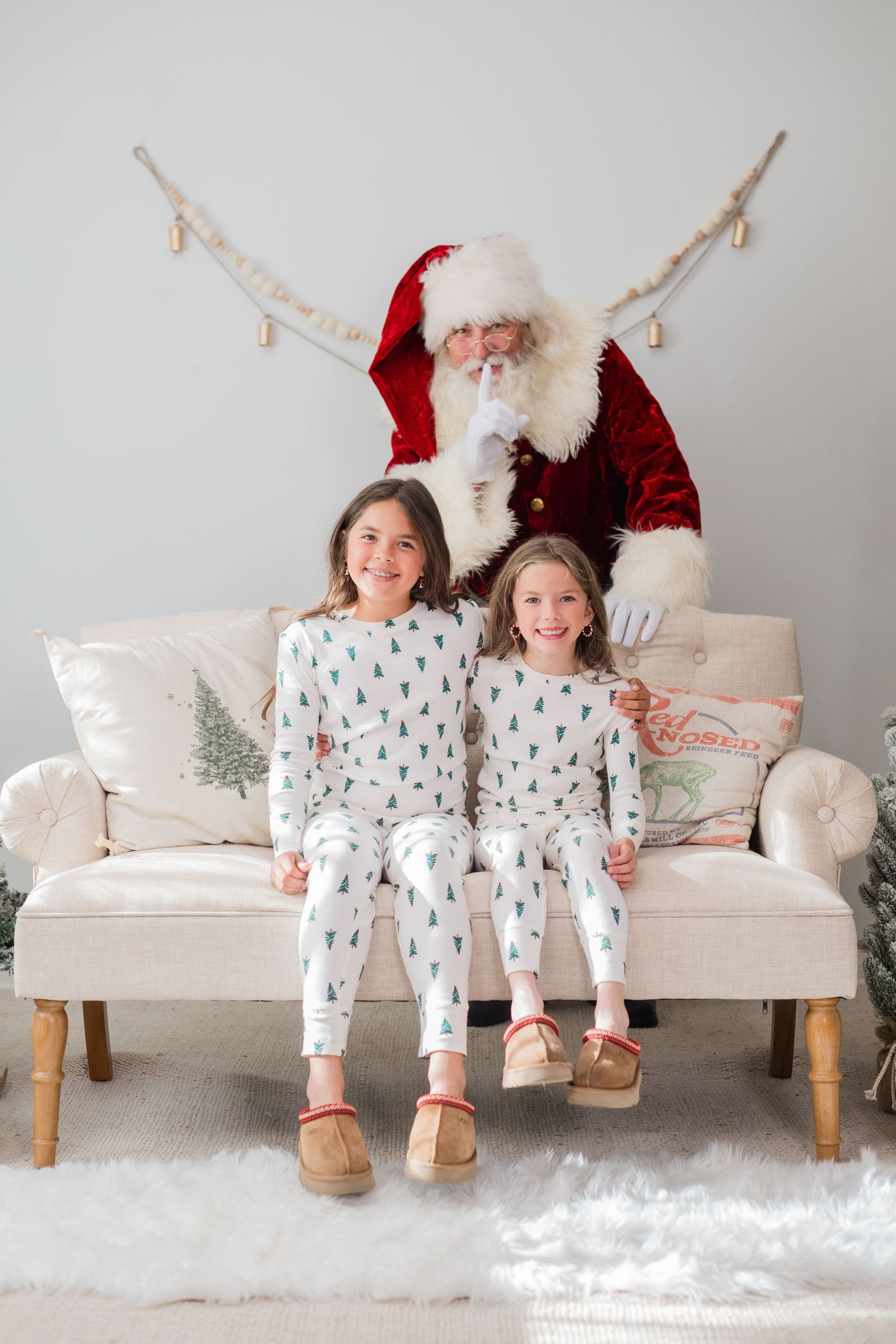 Two girls in matching Christmas tree pajamas sit smiling with Santa Claus on a cream couch in a festive holiday setting.