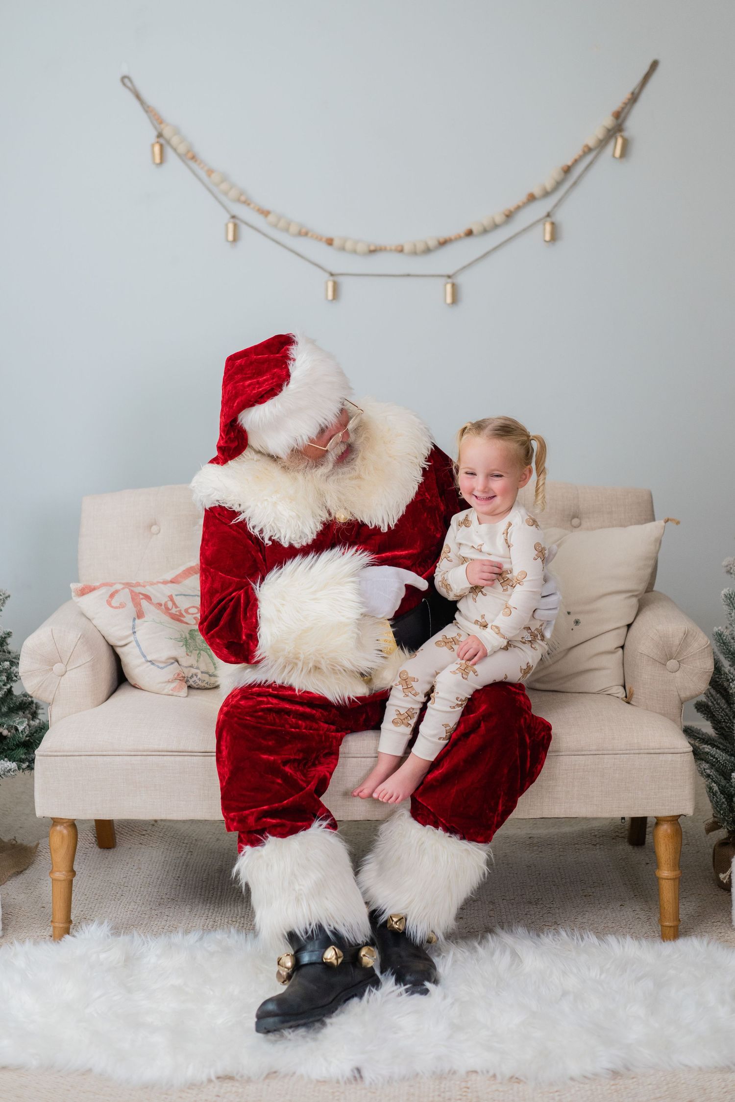 A cheerful toddler sits on Santa Claus's lap on a beige sofa during a festive Christmas photo session.