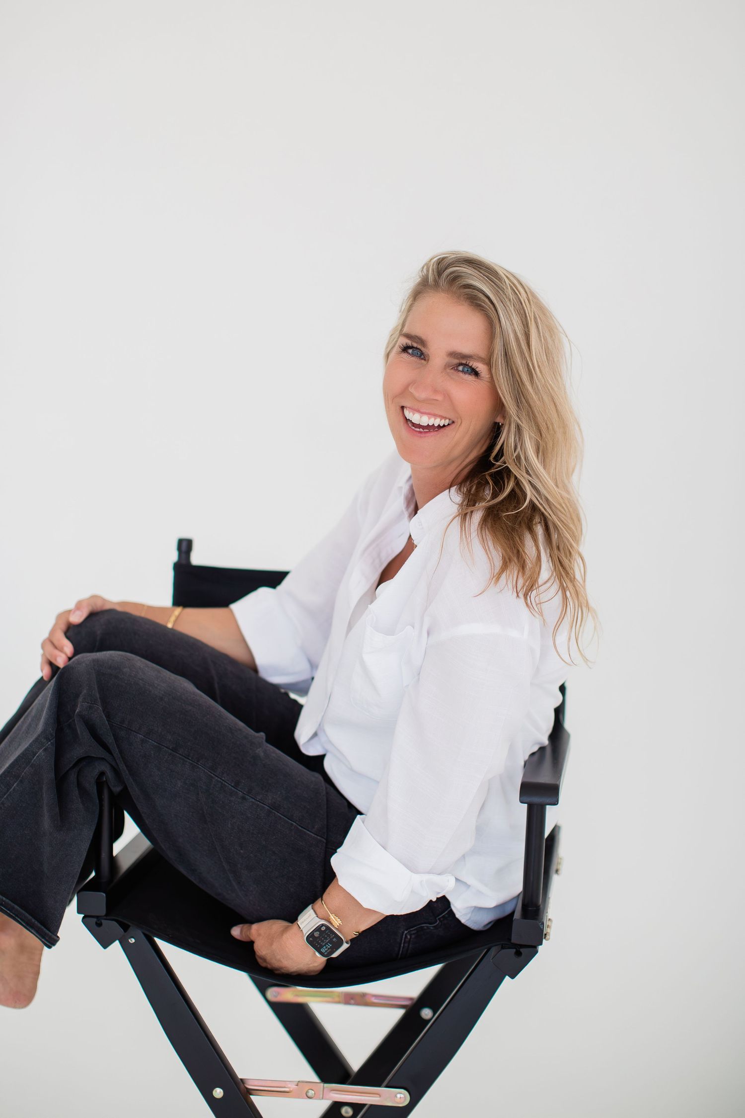Smiling blonde woman in white shirt and dark jeans sitting relaxed in a black director's chair against white background.