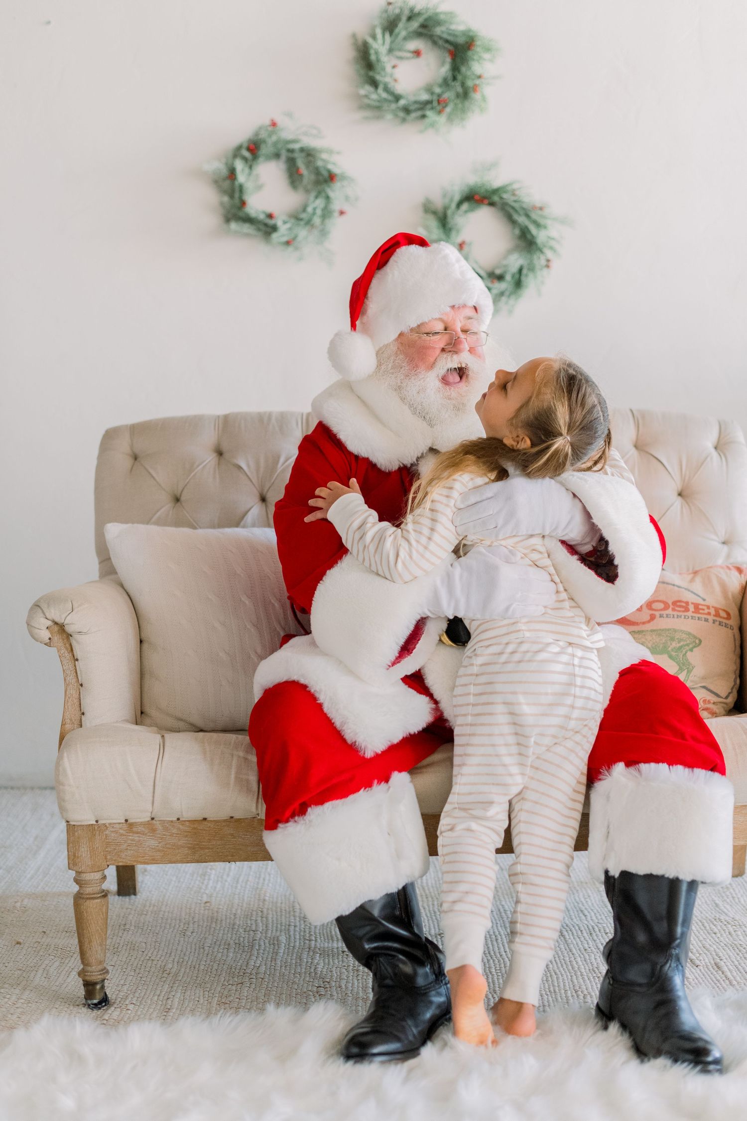 Two children in pajamas hugging Santa Claus sitting on a beige tufted chair with Christmas wreaths on the wall.
