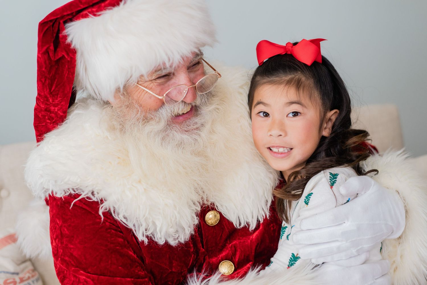 A young girl with a red bow sits smiling with Santa Claus in a classic holiday portrait photo.