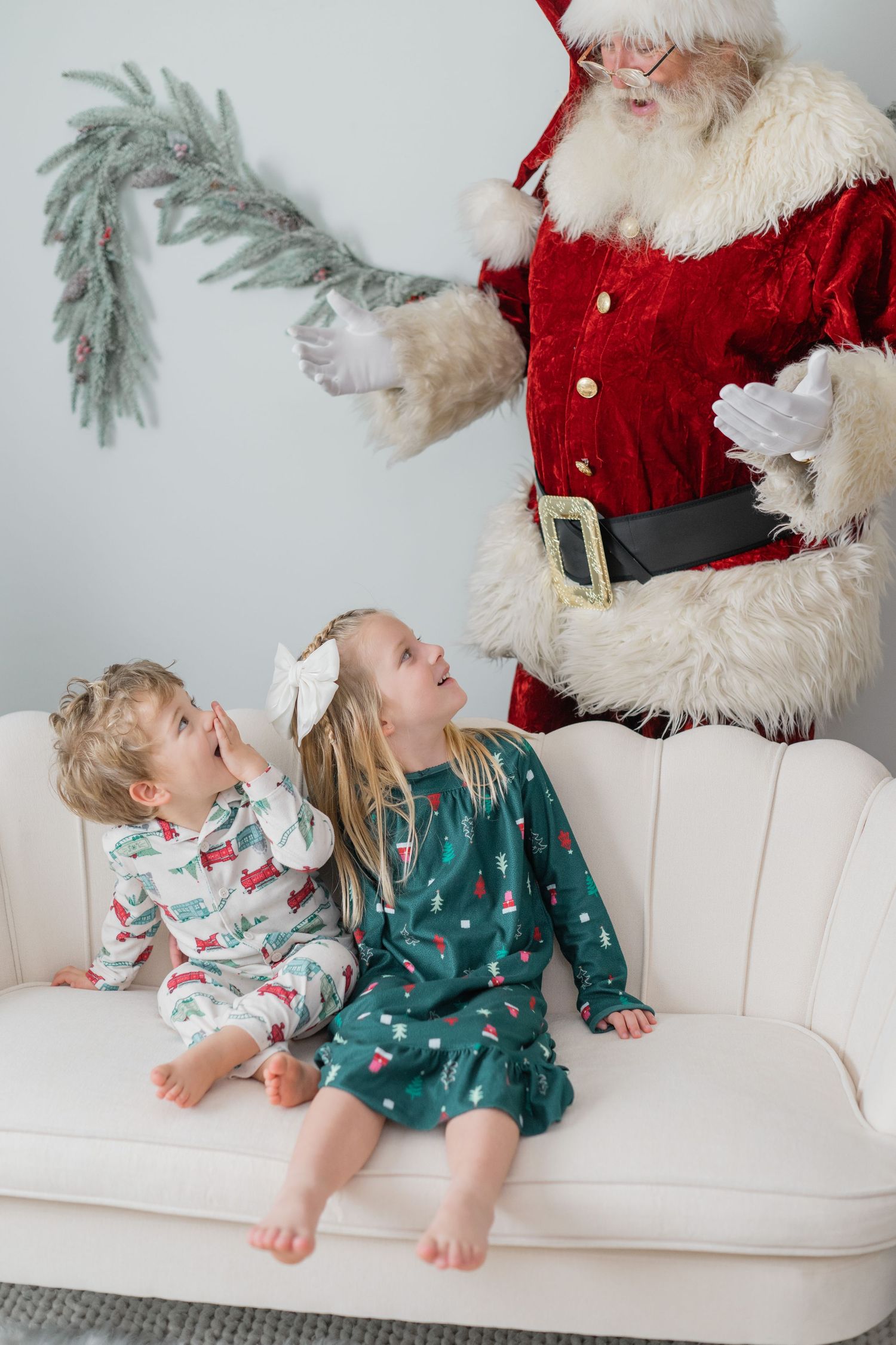 Two young children in Christmas pajamas sit on a white sofa as Santa Claus gestures above them.