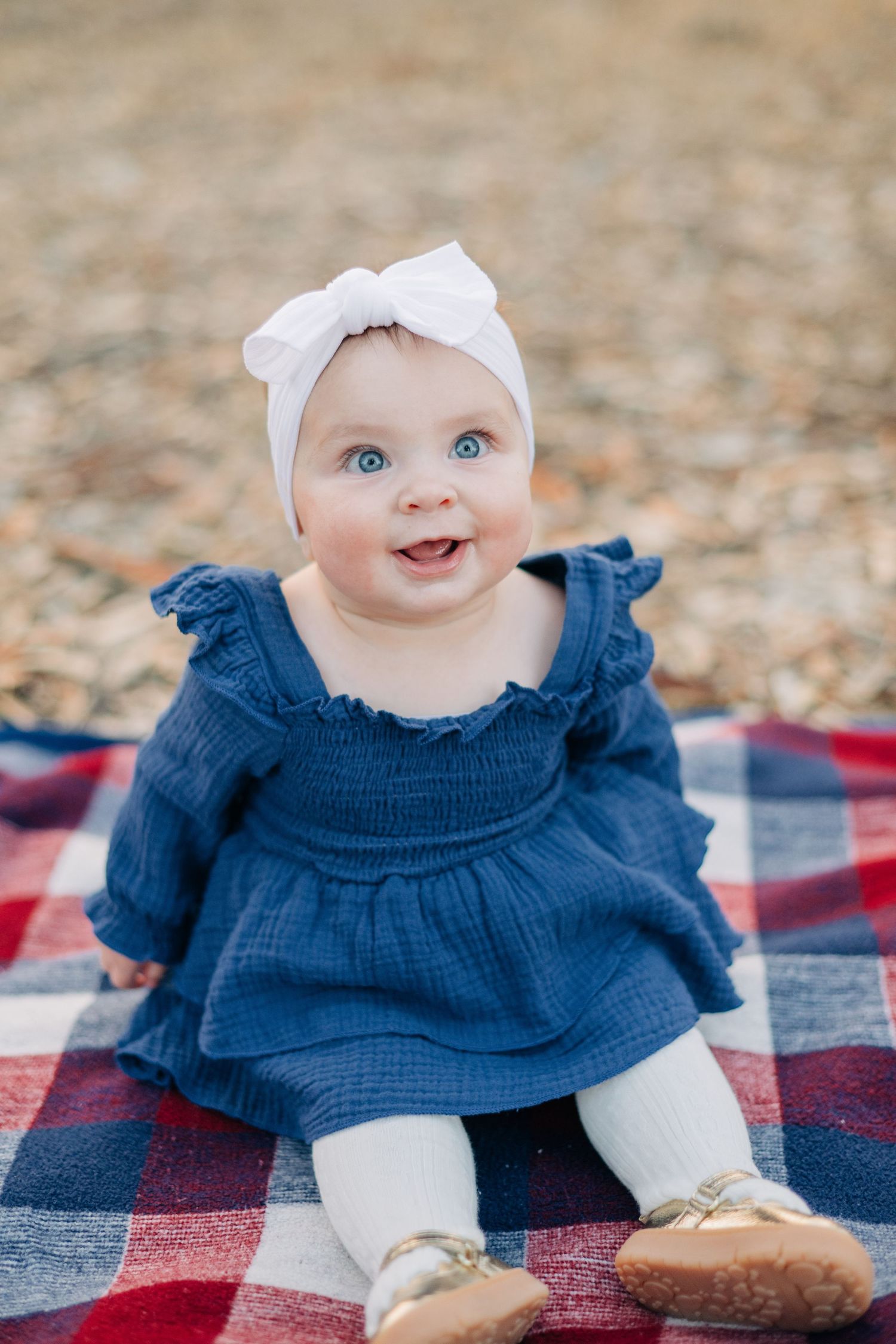 Adorable blue-eyed baby girl in blue dress and white bow headband sitting on plaid blanket outdoors.