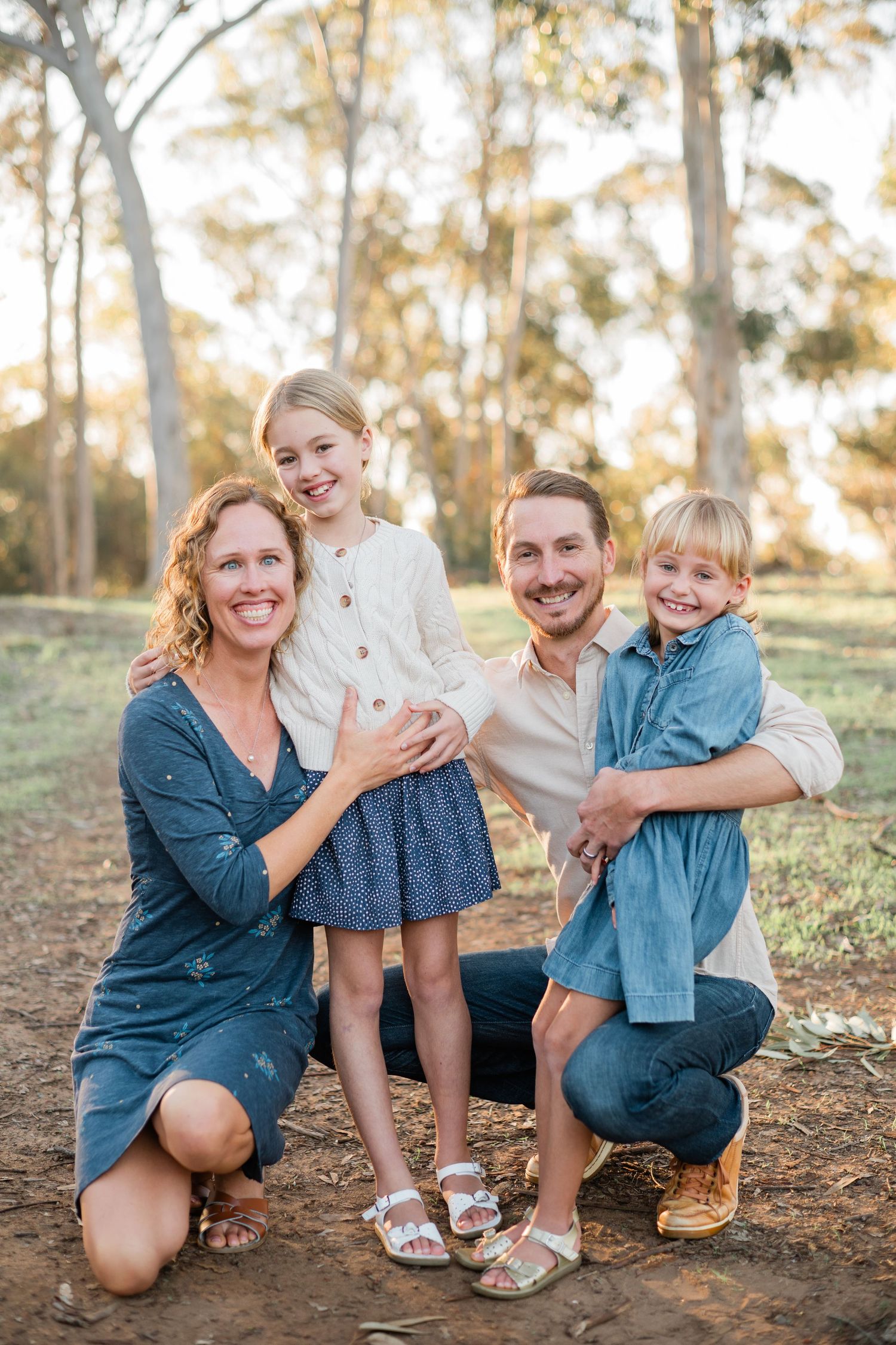 Family of four posing outdoors in golden light among eucalyptus trees, parents kneeling with two smiling young girls.