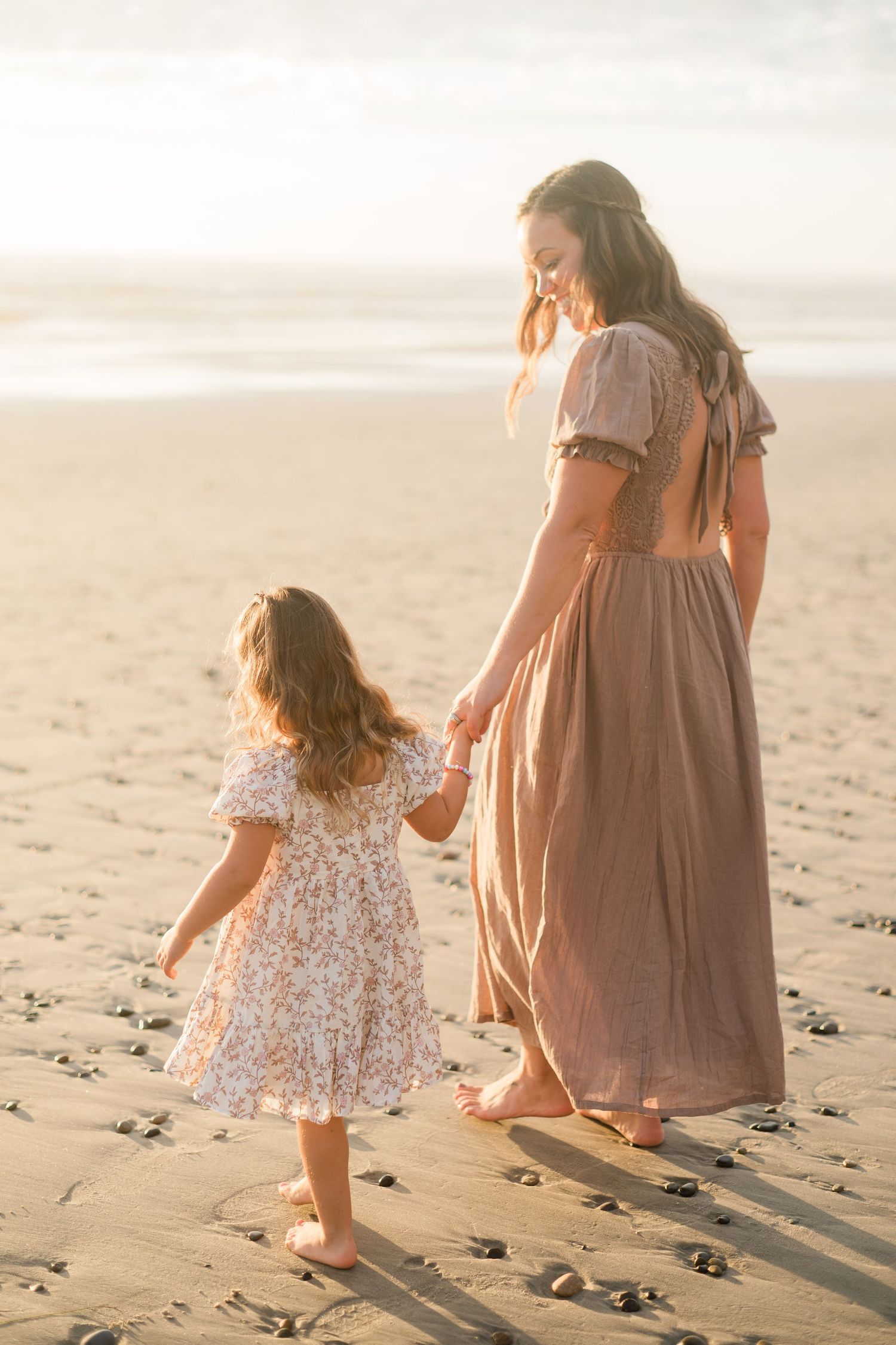 A mother and young daughter in matching dresses hold hands walking on a sunny beach at golden hour.