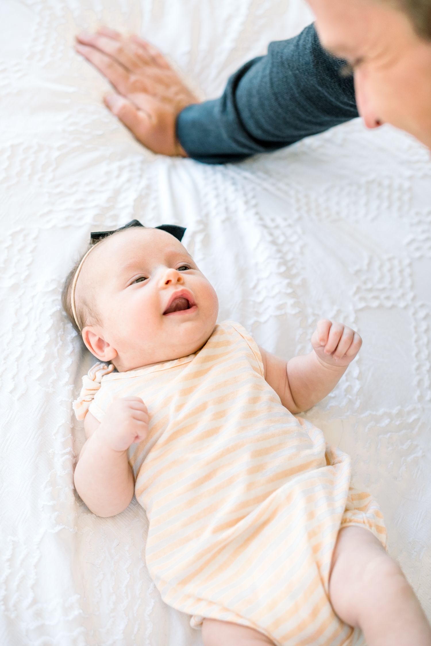 A smiling newborn baby in a striped onesie lies on a white bed while a parent reaches out nearby.
