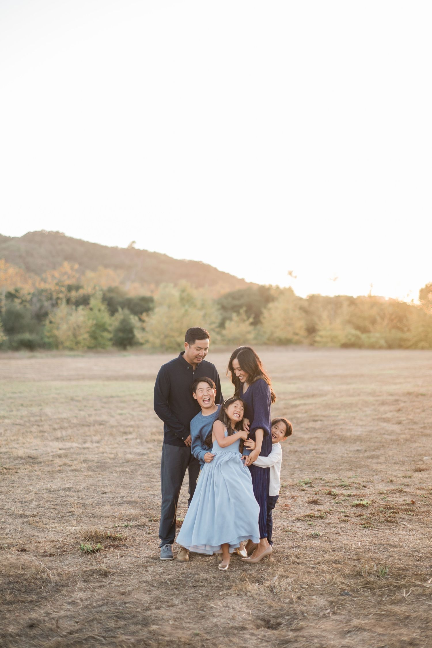 Family of four laughing together in a golden field at sunset, with hills and trees in the background.