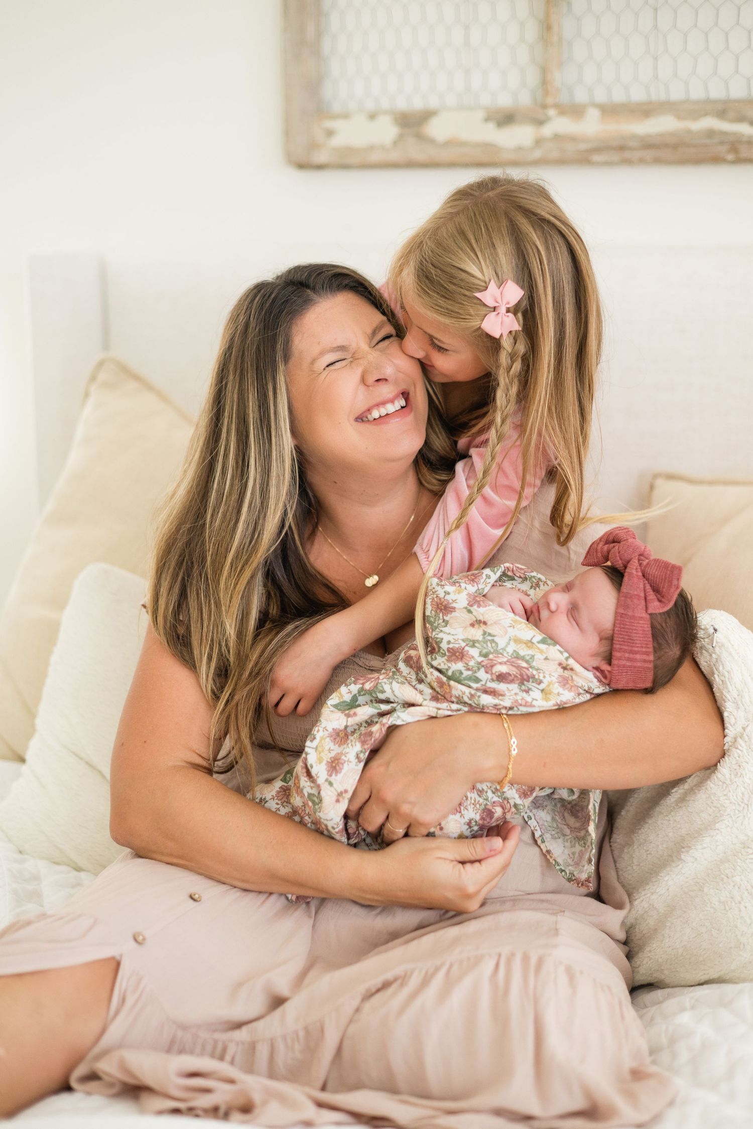Laughing mother holds sleeping newborn while young daughter gives her a kiss on the cheek on a bed.
