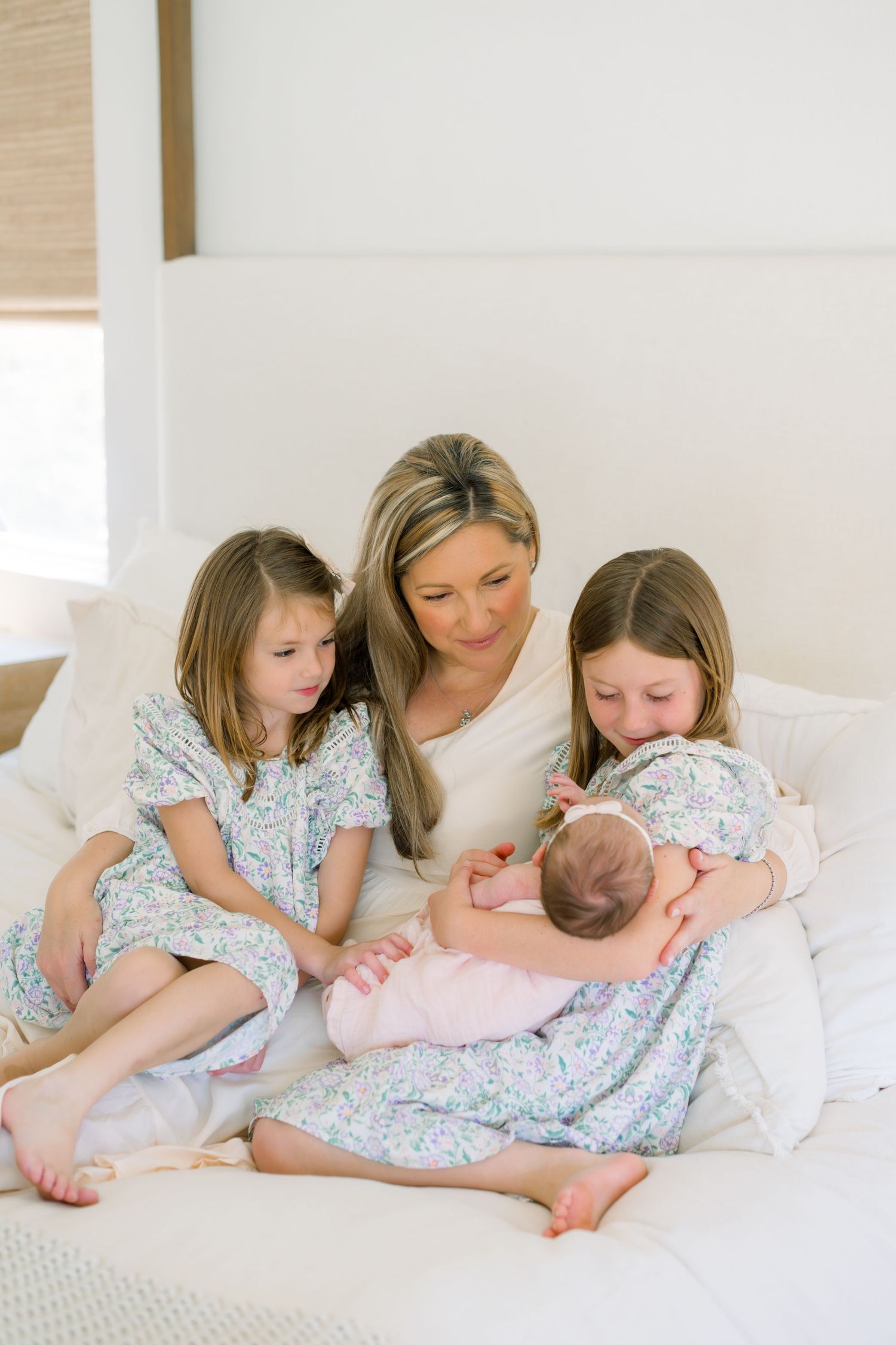 A mother sits on a white bed with her two young daughters in floral dresses, lovingly gazing at their newborn baby sister.