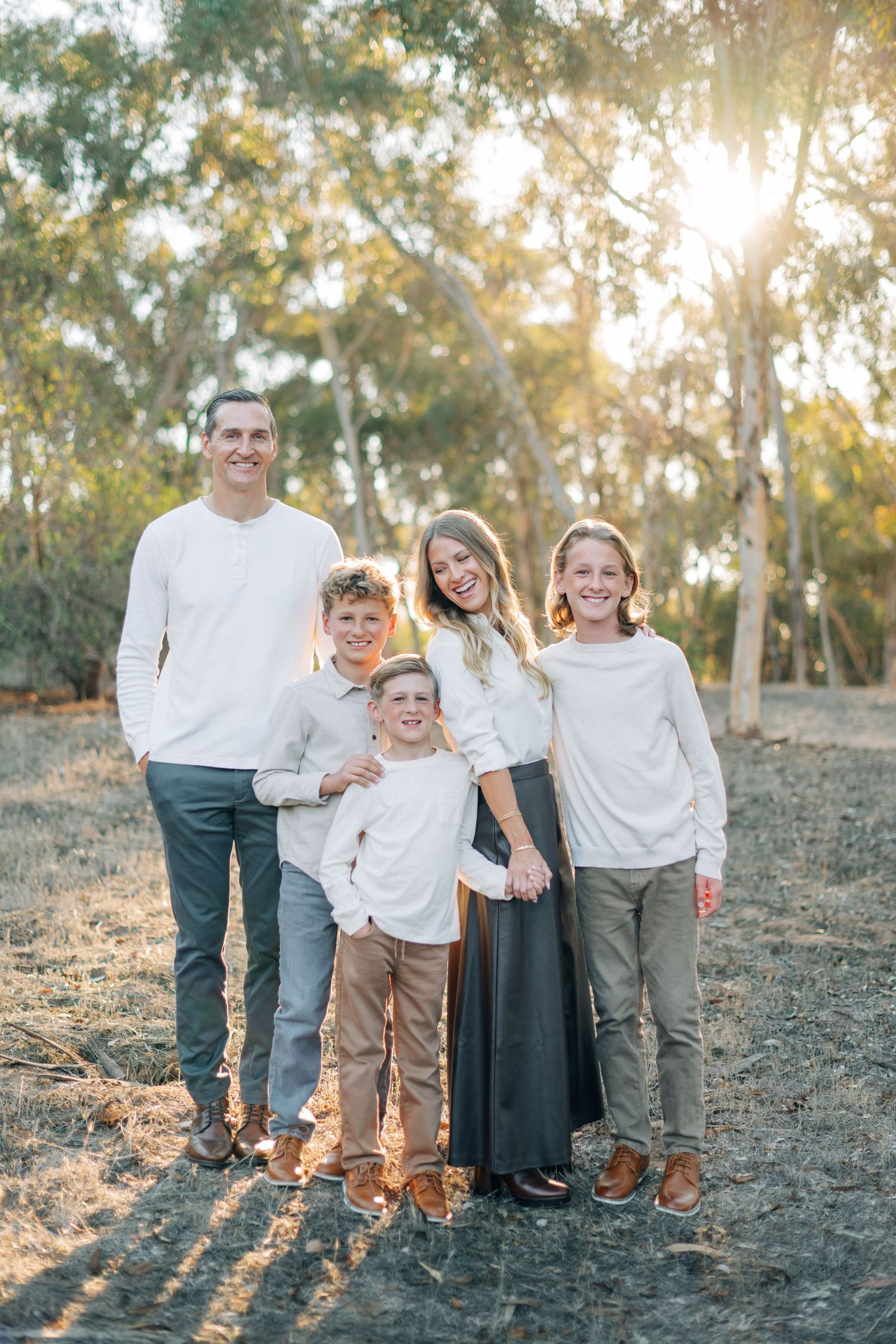 Family of five posing outdoors in white tops among eucalyptus trees in warm golden sunlight.