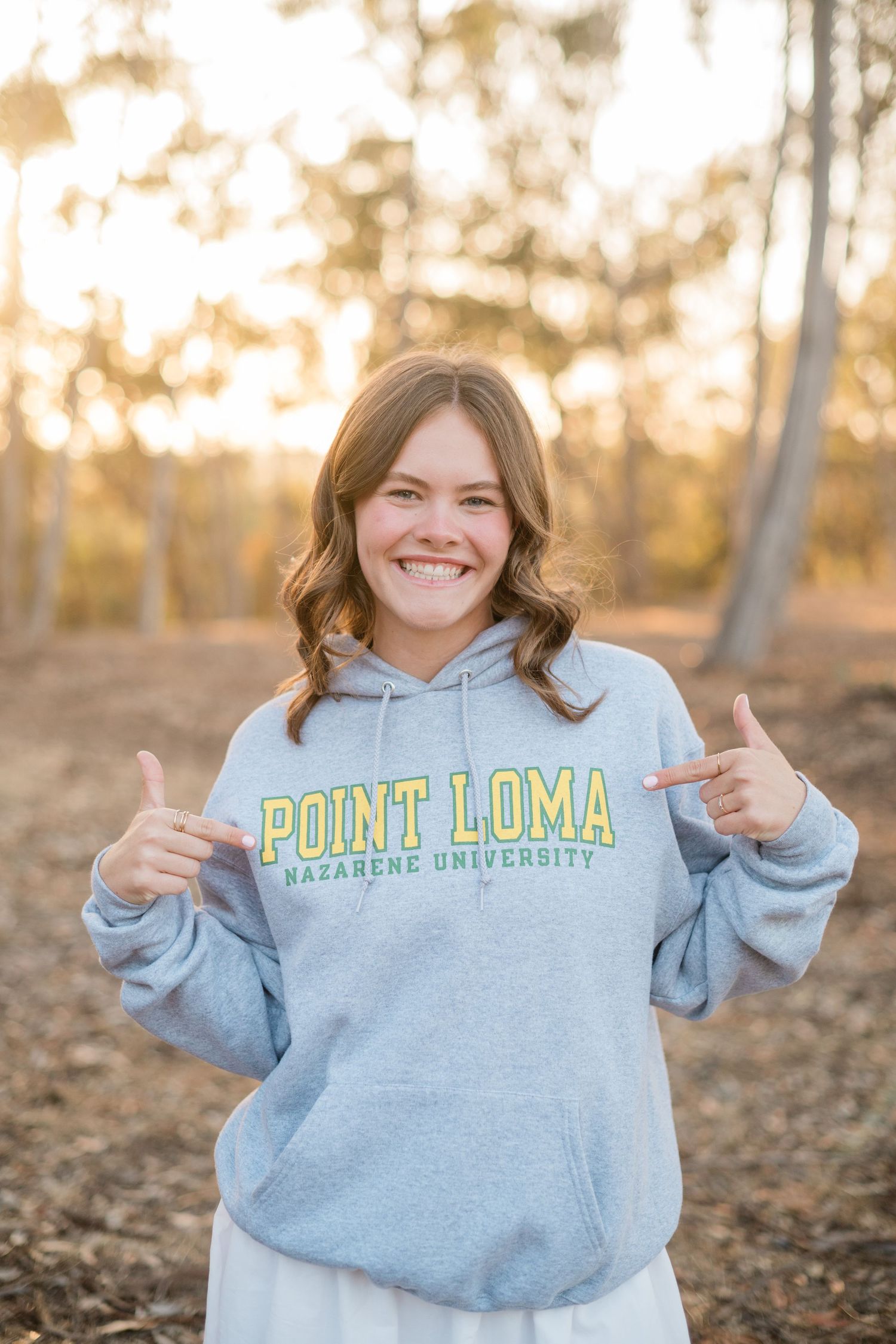 Smiling young woman pointing at her gray Point Loma Nazarene University hoodie outdoors at golden hour.