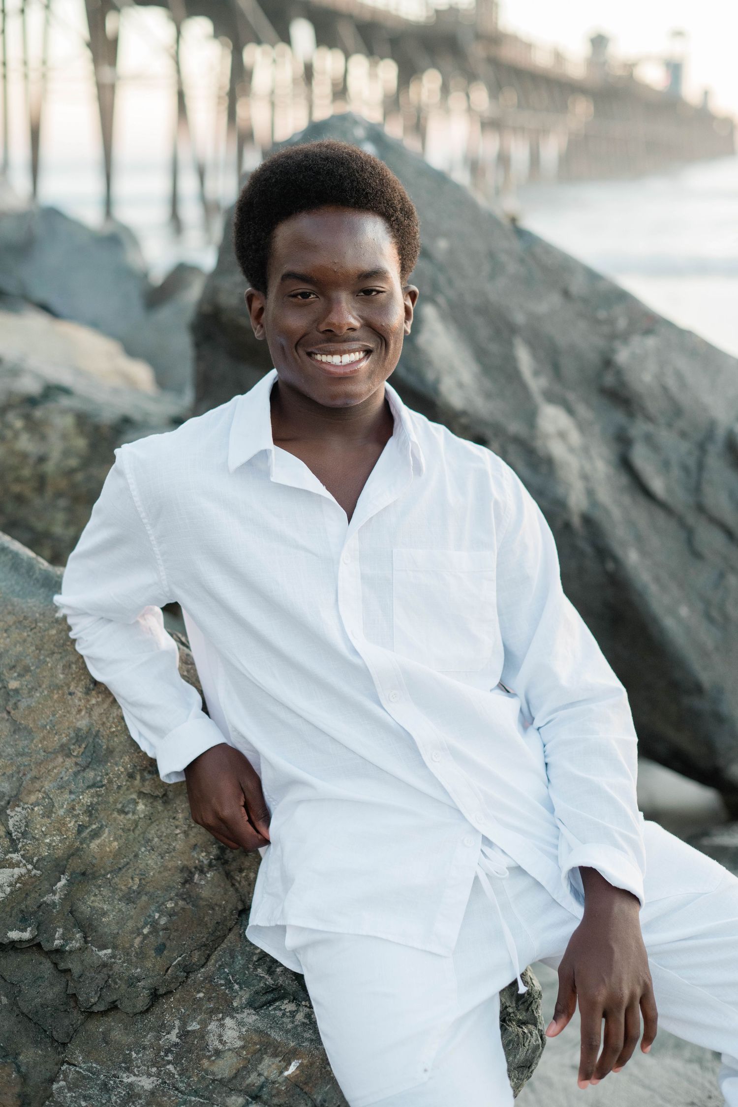 Smiling young Black man in white outfit sitting on rocks near a pier at the beach.