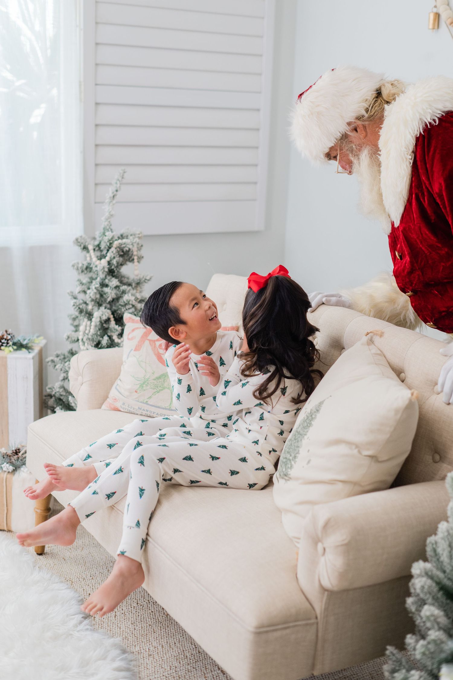 Two children in Christmas tree pajamas sit on a couch smiling at Santa Claus in a festive holiday setting.