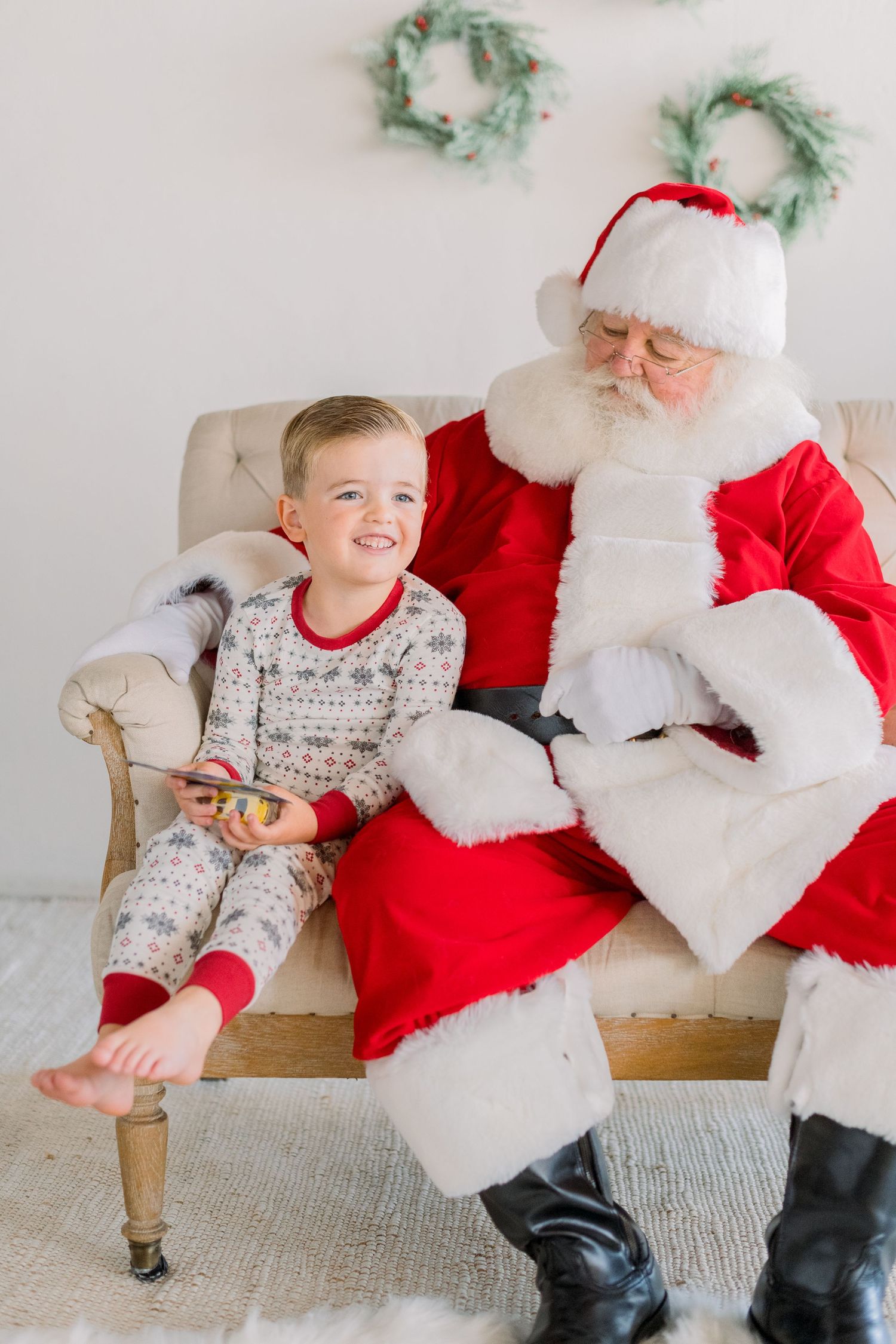 A smiling toddler boy in holiday pajamas sits with Santa Claus on a beige chair during a Christmas photo session.
