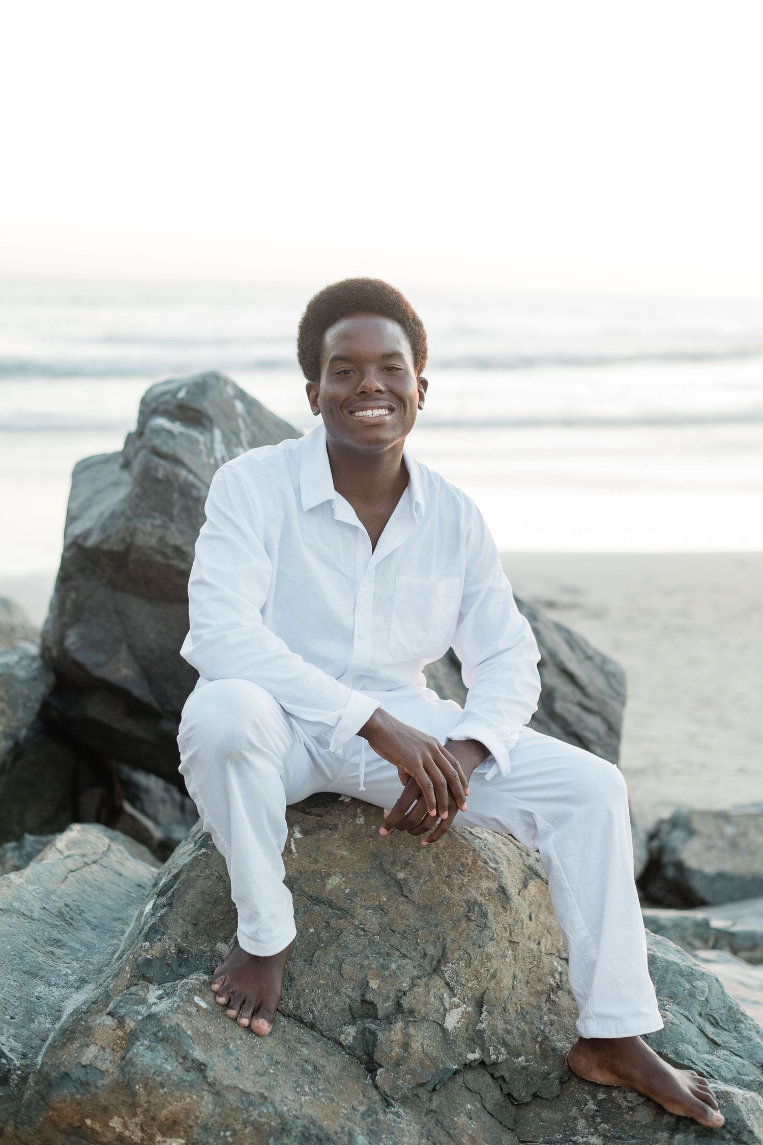 Smiling young man in white outfit sitting barefoot on rocks at a beach with ocean waves in the background.