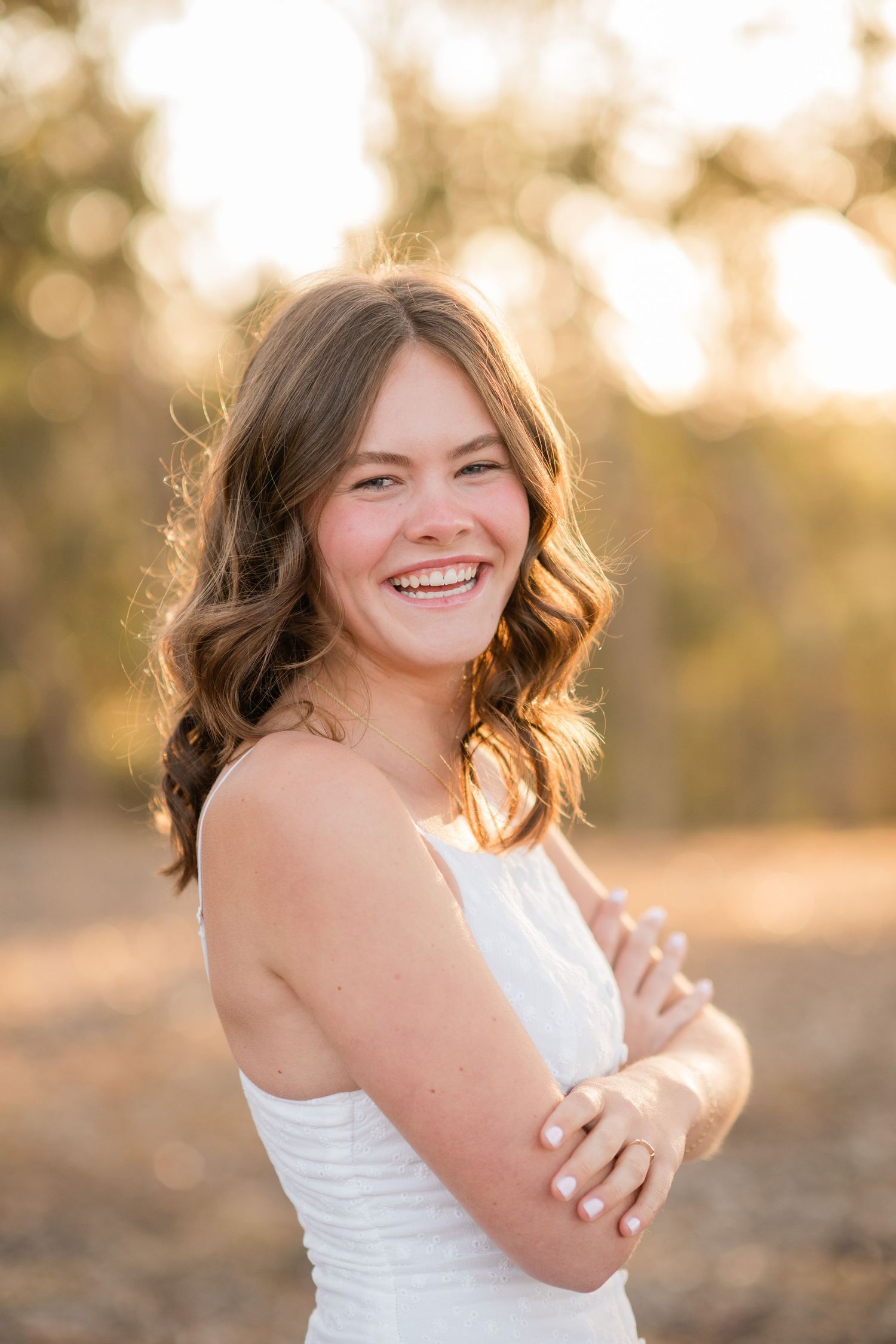 Young woman in white dress smiling outdoors in golden sunlight with wavy brown hair.