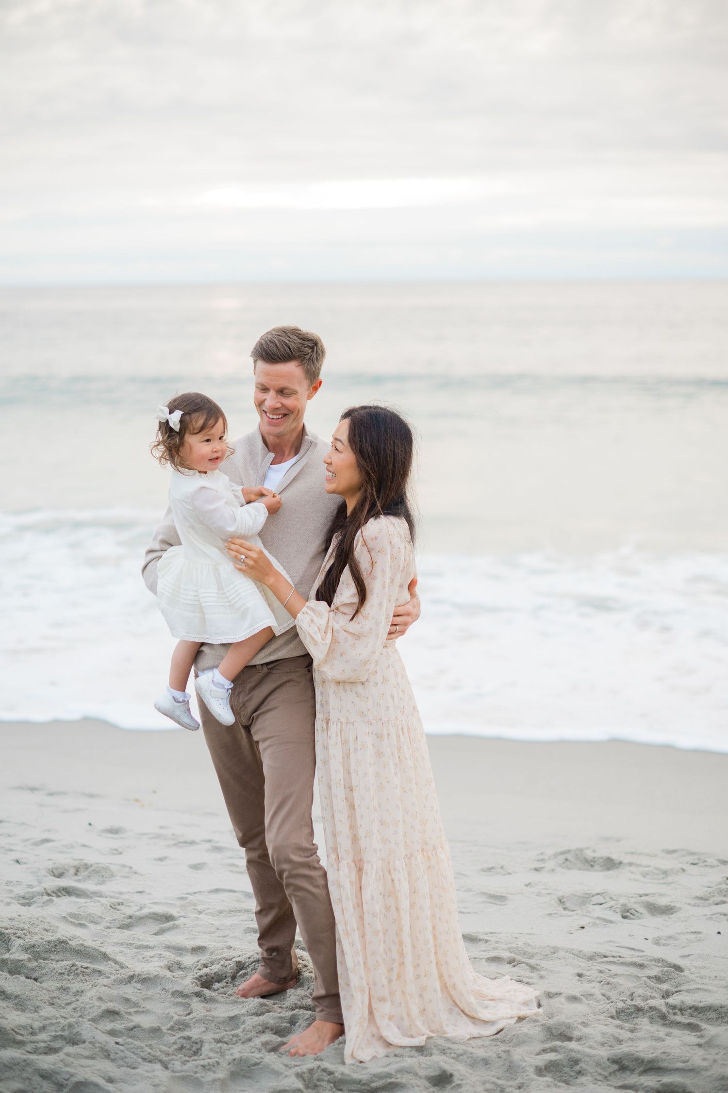 Happy family of three poses on a sandy beach, parents smiling at their toddler daughter in white dress.