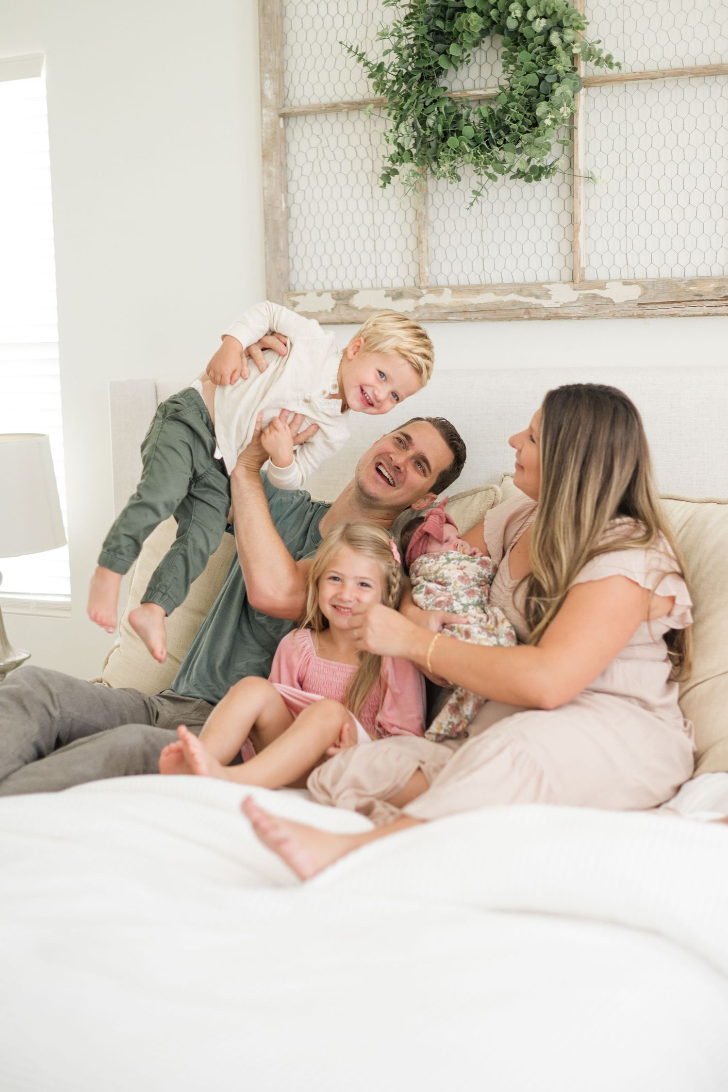 Happy family of five laughing and playing together on a bed in a bright farmhouse-style bedroom.