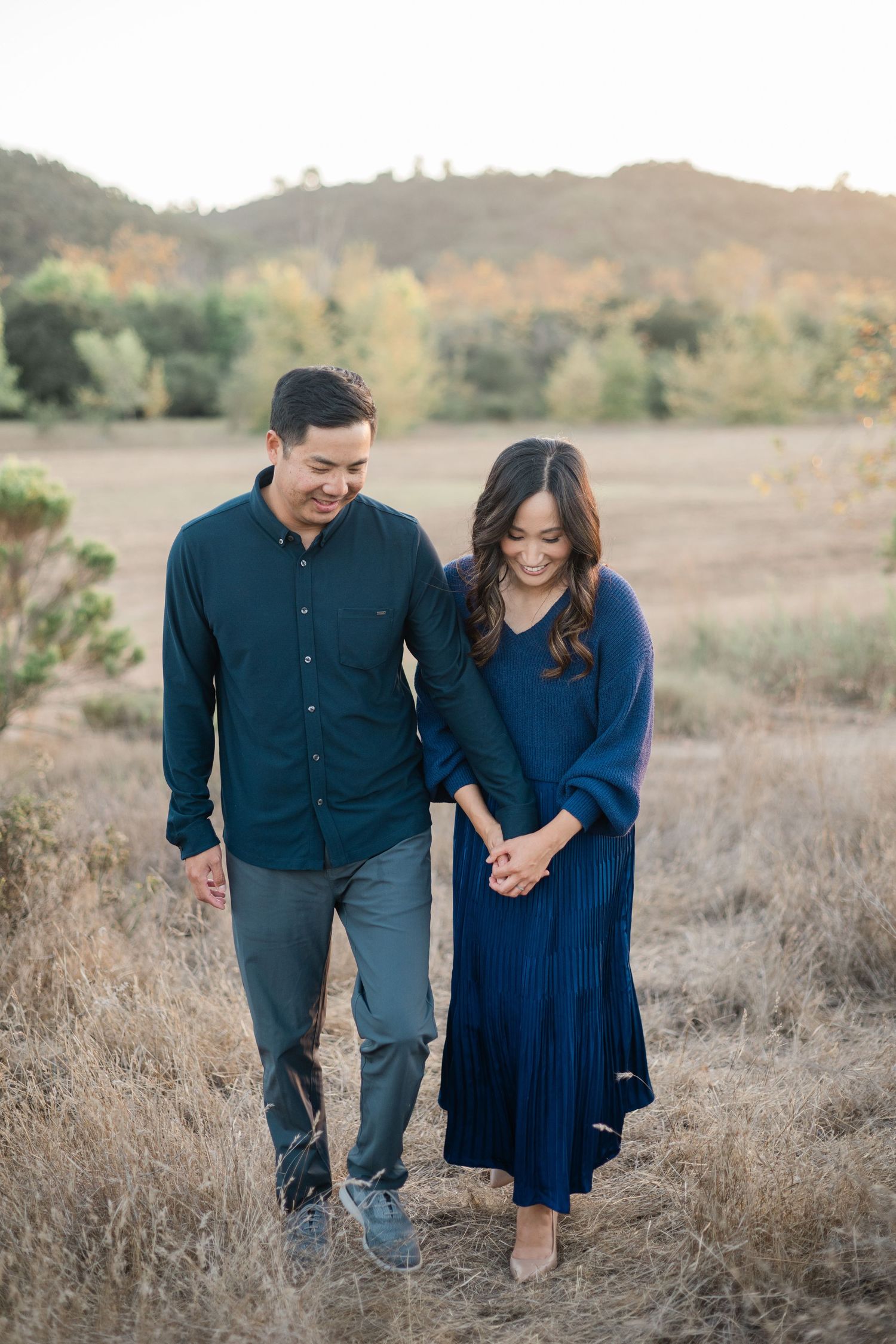 A smiling Asian couple walks hand in hand through a golden field at sunset wearing navy and teal outfits.