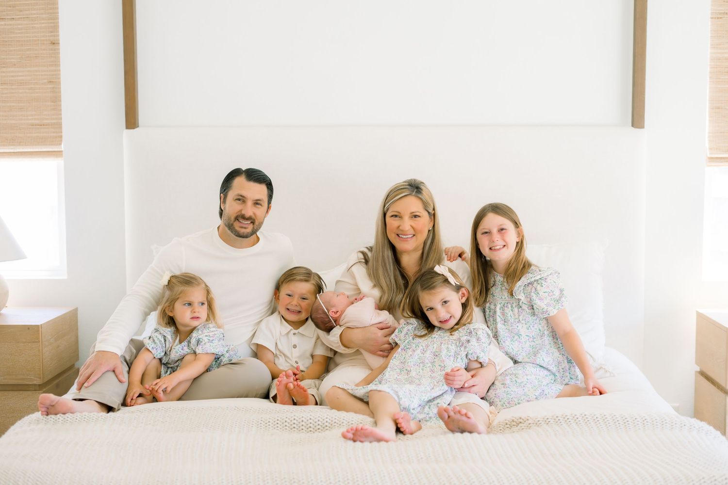A happy family of seven poses together on a white bed, smiling warmly in coordinated light-colored outfits.