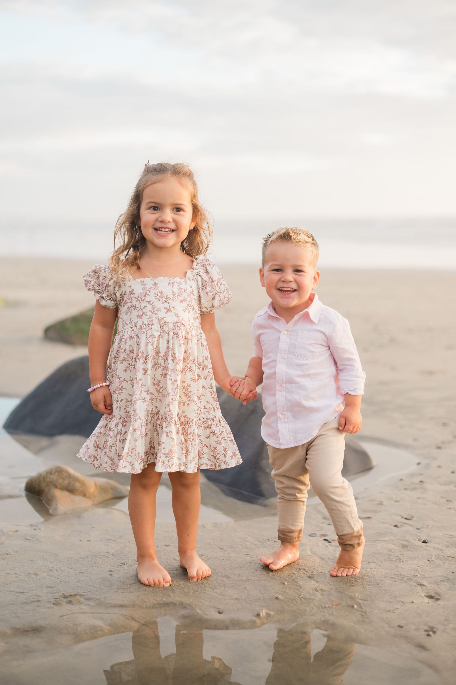 Two young children holding hands and smiling on a sandy beach, girl in floral dress and boy in pink shirt.