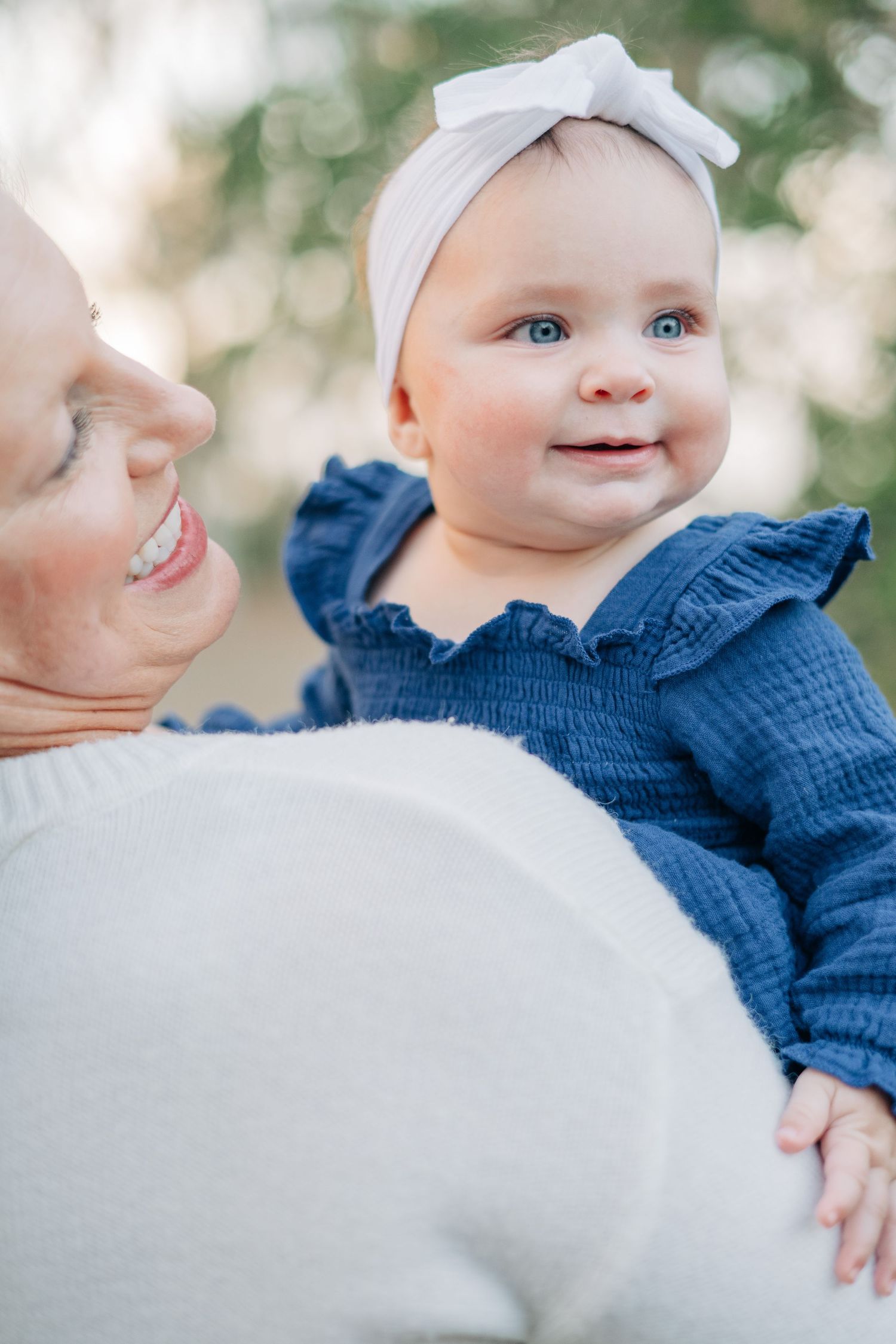Smiling baby girl in blue ruffle dress and white bow headband being held by a laughing woman outdoors.