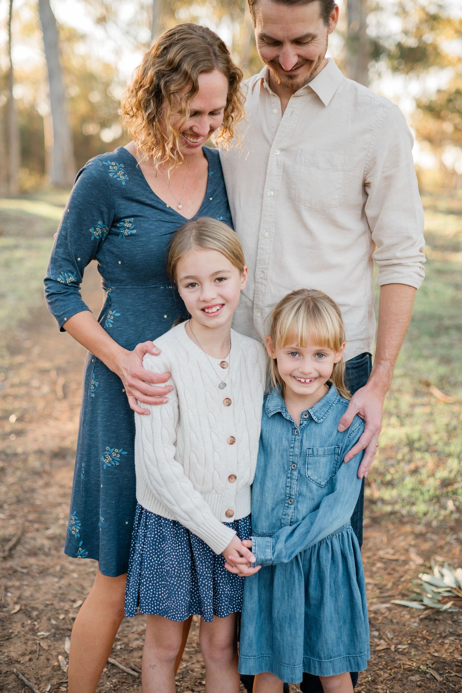 Family of four poses outdoors, parents smiling at two blonde girls in blue dresses among trees.