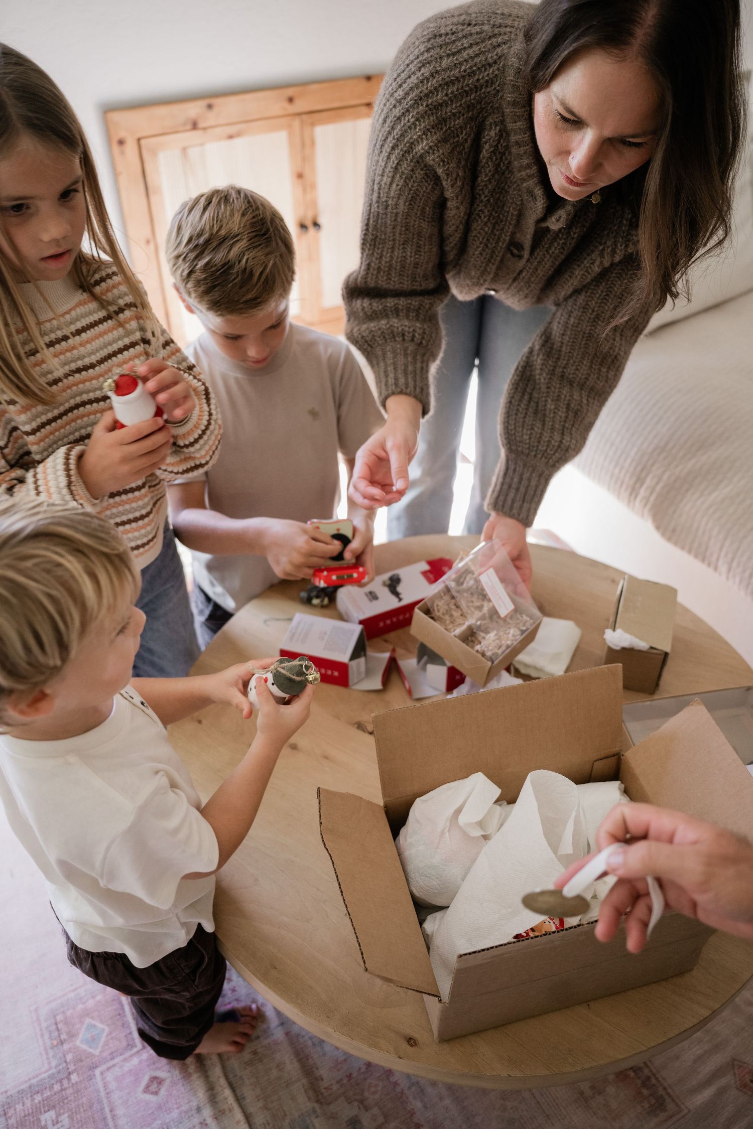 Family unpacking Christmas decorations from a cardboard box together around a wooden table in a cozy living room.