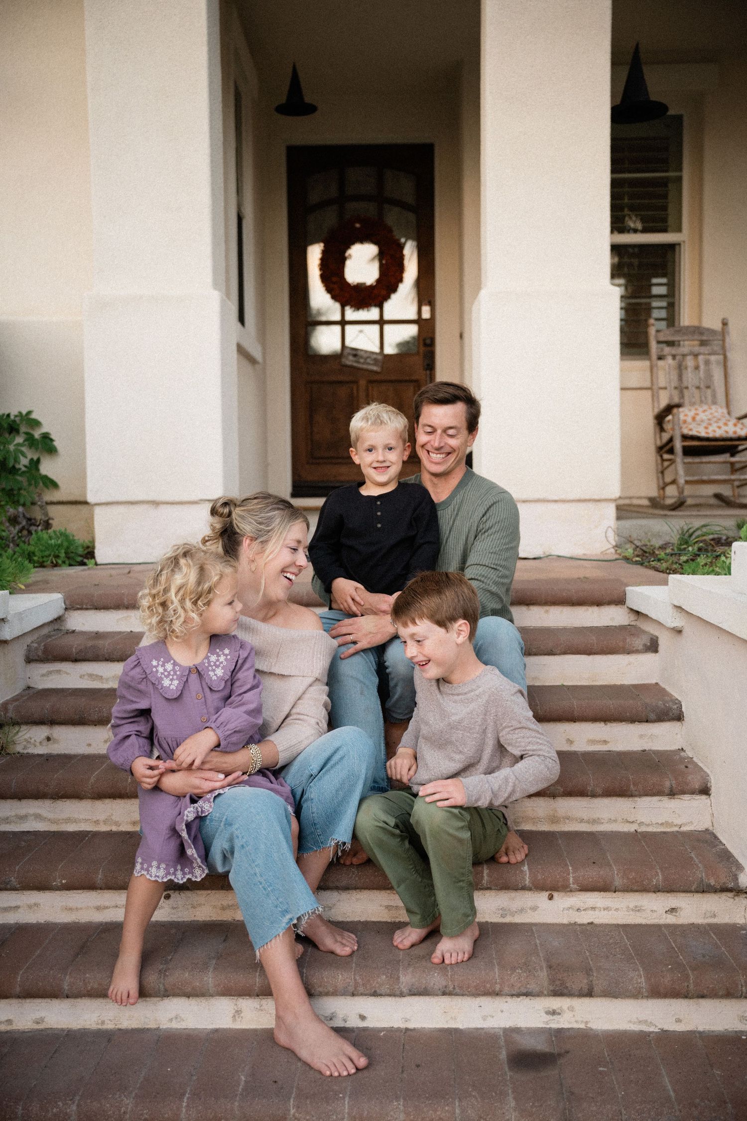 Happy family of five sitting barefoot on front porch steps of a white home with a wreath on door.