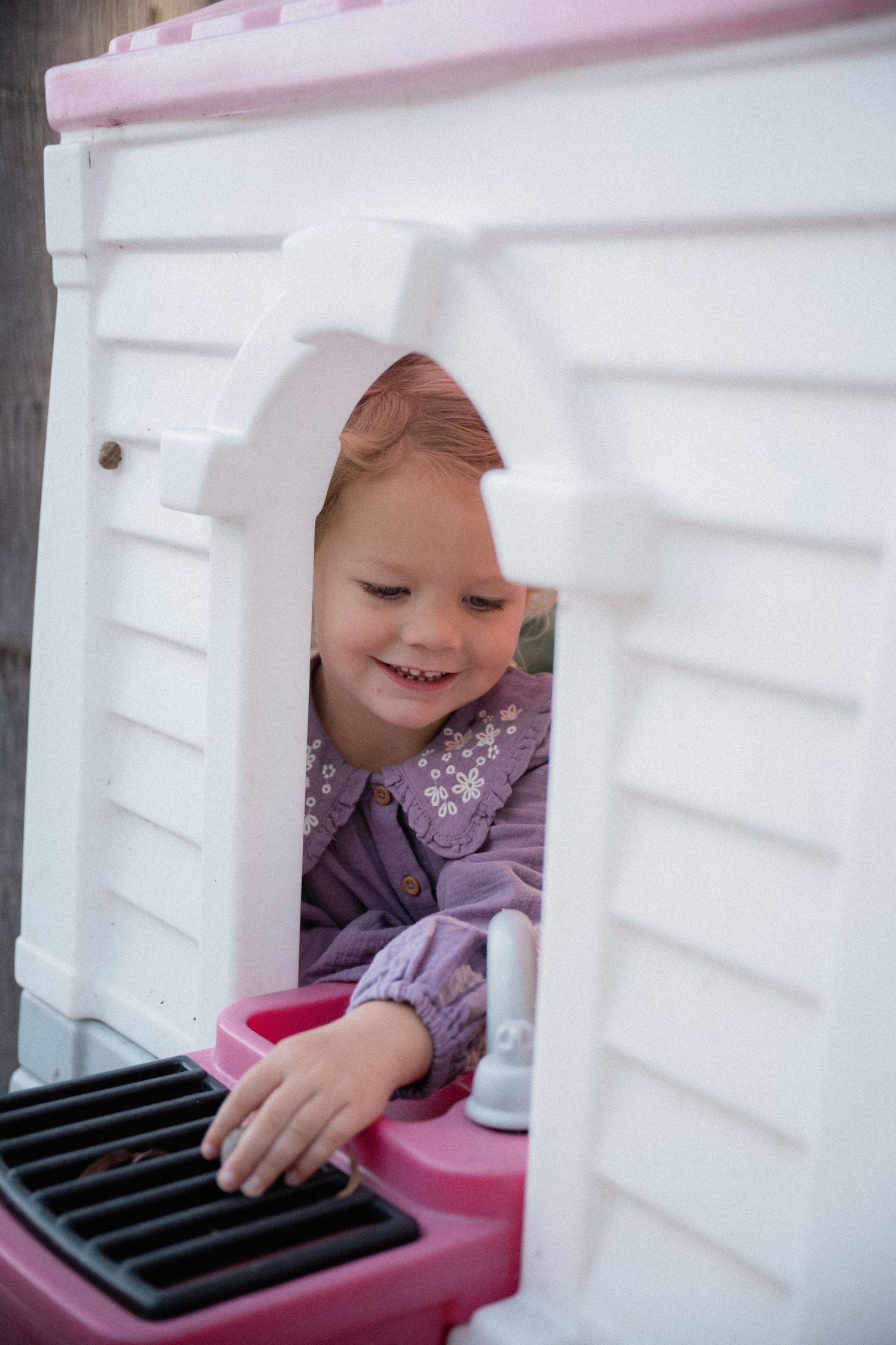 Smiling toddler girl in purple dress playing inside a pink and white plastic playhouse outdoors.