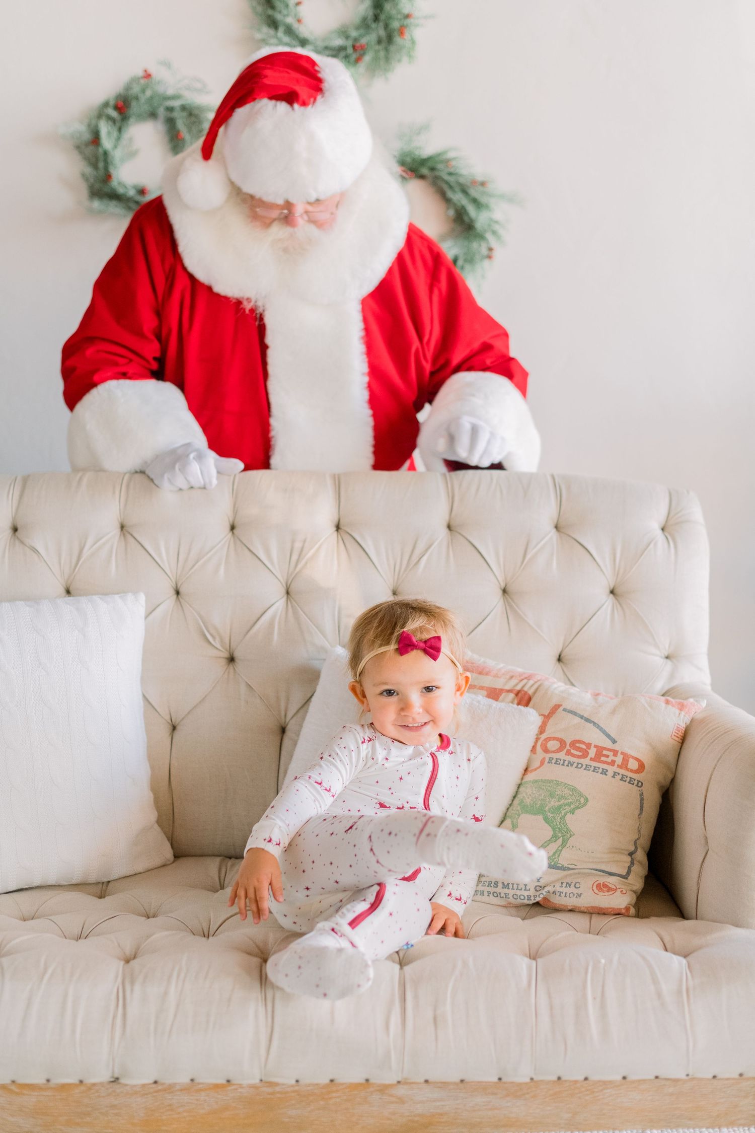 Smiling baby girl in white pajamas sits on a cream tufted sofa with Santa Claus behind her at Christmas.